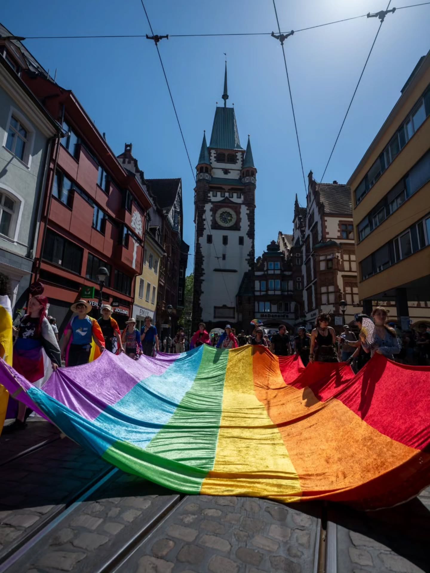 Menschen halten eine große Regenbogenfahne auf einer Straße mit Straßenbahnschienen, im Hintergrund ein markanter Uhrturm und historische Gebäude unter blauem Himmel. (KI-Alternativtext)