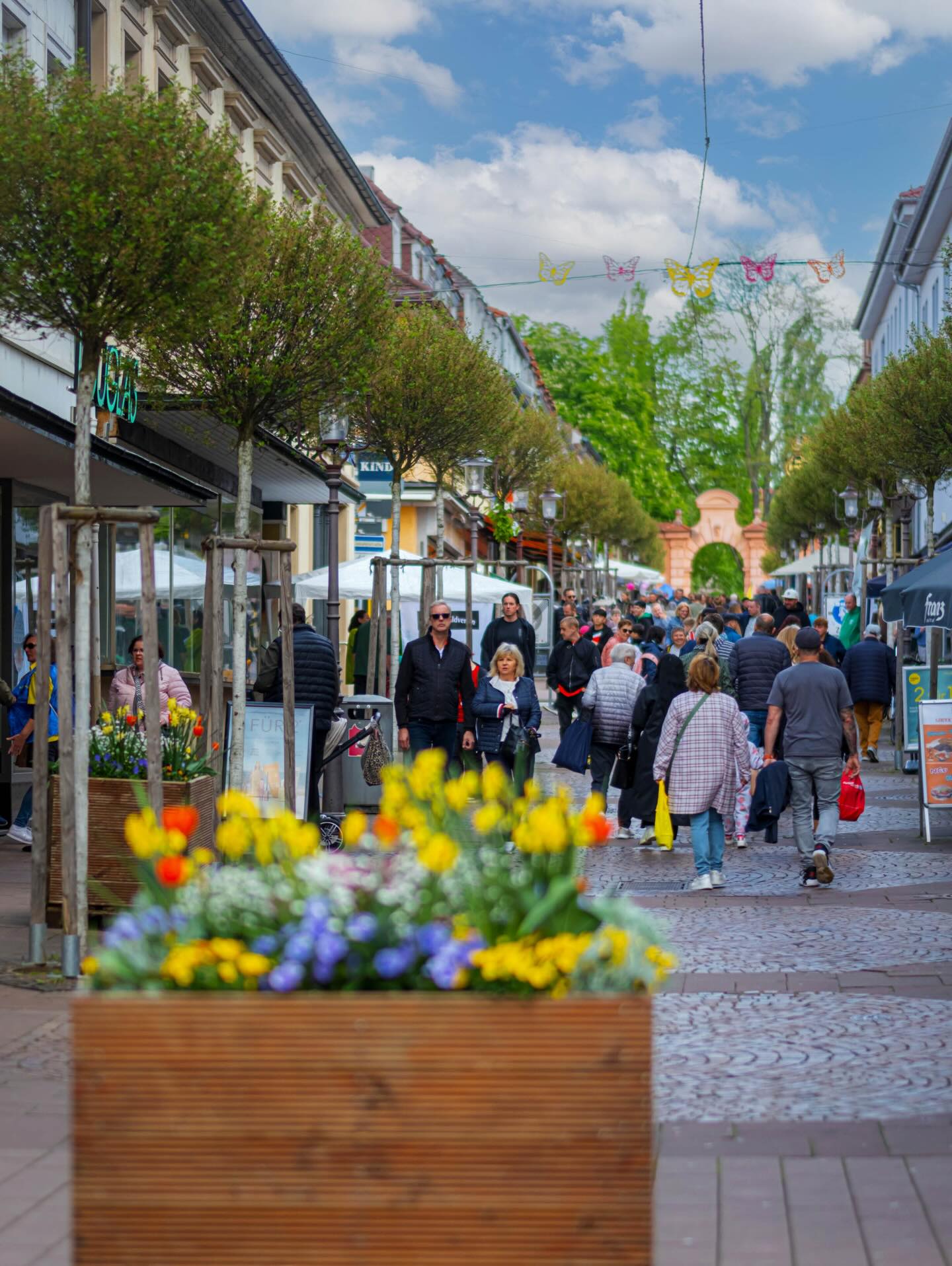 Dieser Alternativtext wurde KI-generiert: Eine belebte Fußgängerzone mit Geschäften und Bäumen. Viele Personen sind auf dem Bürgersteig unterwegs, einige tragen bunte Kleidung und es gibt Blumenschmuck im Vordergrund.