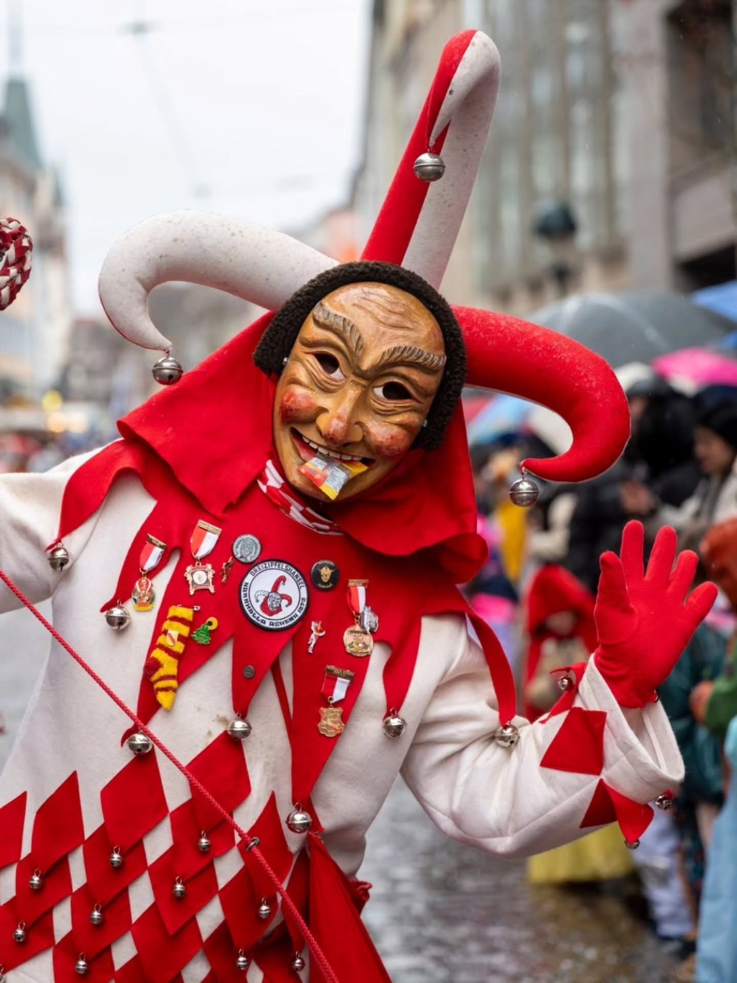 Dieser Alternativtext wurde KI-generiert: Ein Kostüm mit einer auffälligen Maske und Glocken ist zu sehen. Das Outfit ist rot und weiß kariert und mit verschiedenen Abzeichen verziert.