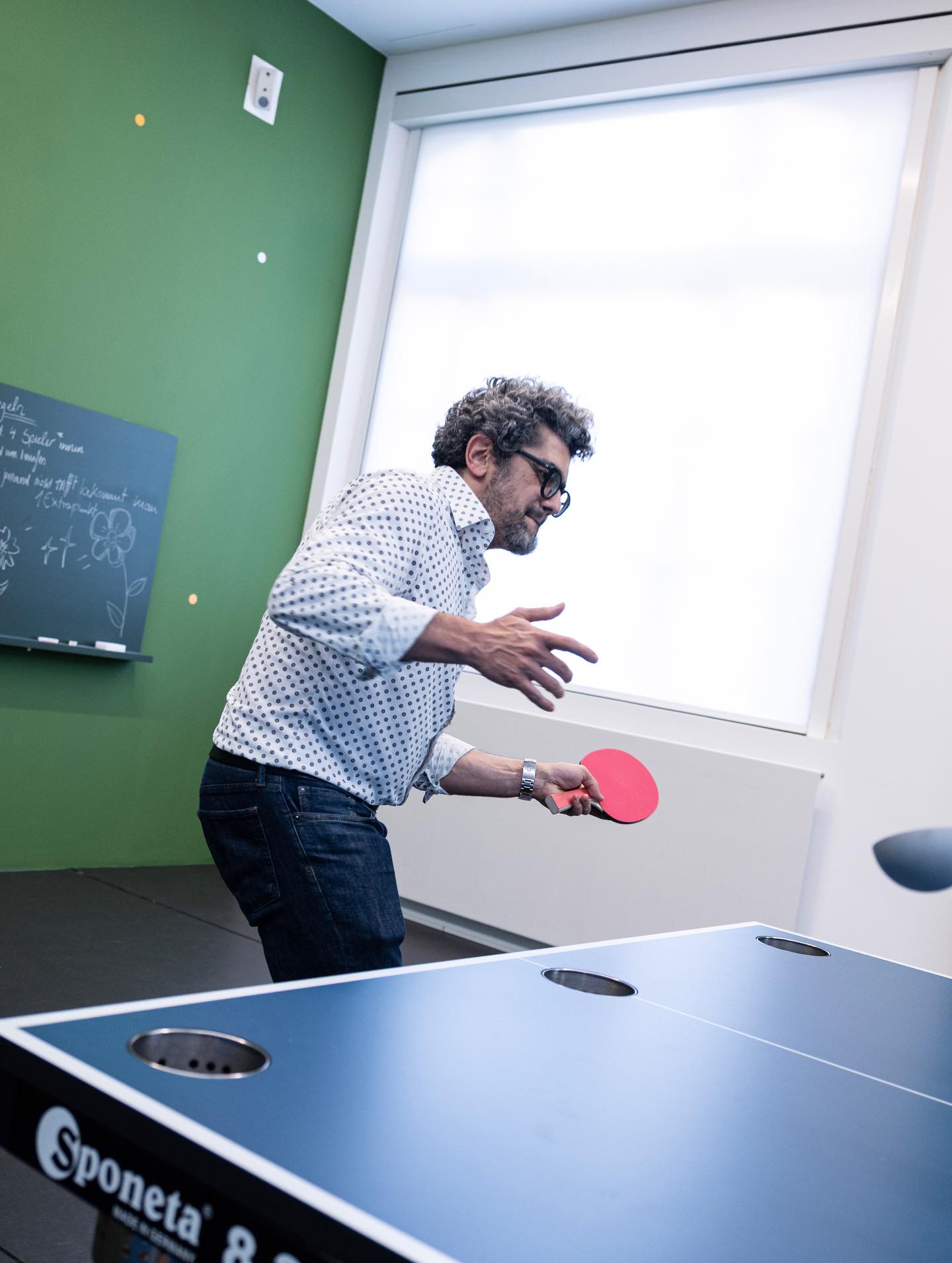 Dieser Alternativtext wurde KI-generiert: Ein Mensch spielt Tischtennis in einem Raum mit grünen Wänden und einer Tafel. Das Bild ist farbig und zeigt eine dynamische Szene.