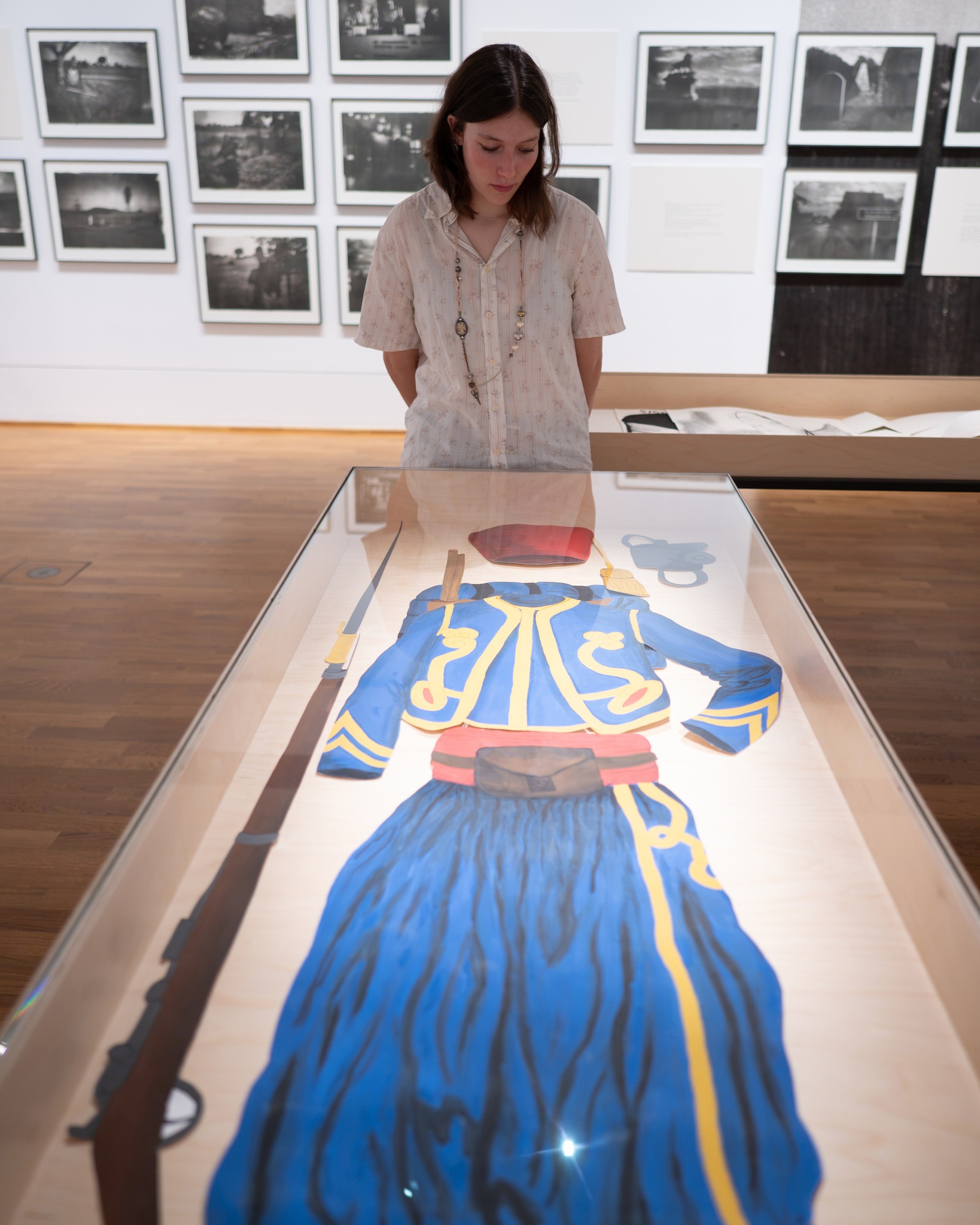 Eine Person betrachtet in einer Glasvitrine ein flach ausgelegtes, blaues Kostüm mit gelben Verzierungen, einem roten Hut und einem Schwert. Im Hintergrund hängen an der Wand zahlreiche gerahmte Schwarz-Weiß-Fotografien. (KI-Alternativtext)