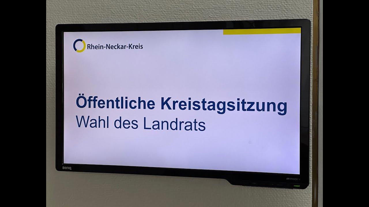 Dieser Alternativtext wurde KI-generiert: Ein Bildschirm zeigt eine Präsentation mit Text in deutscher Sprache. Der Titel der Präsentation bezieht sich auf eine öffentliche Sitzung und die Wahl eines Landrats.