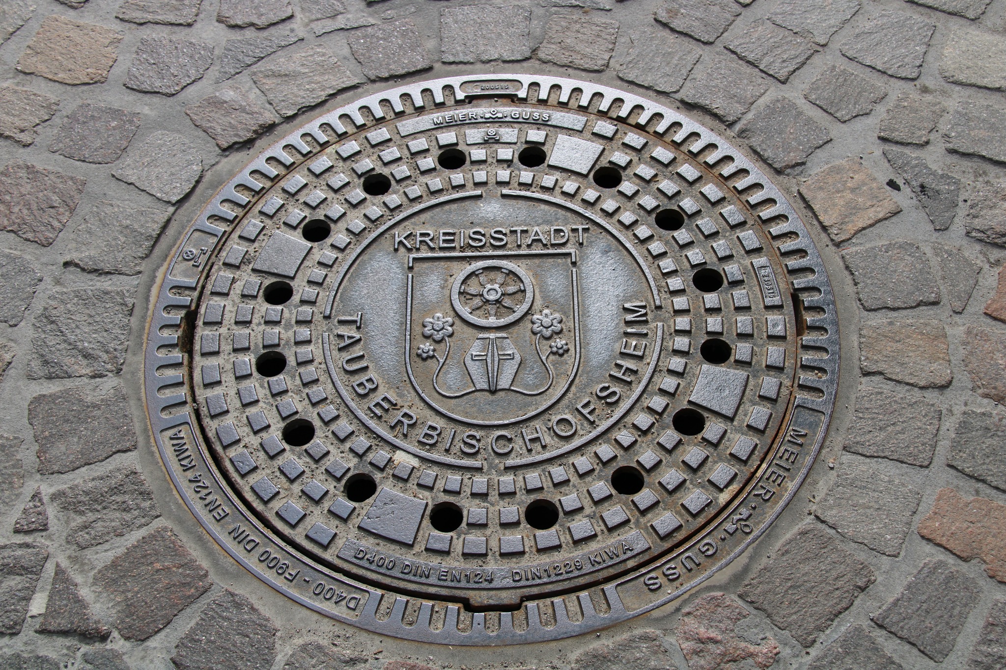 Dieser Alternativtext wurde KI-generiert: Ein runder, grauer Gullydeckel ist in einen Pflastersteinboden eingelassen. Der Deckel zeigt einen Wappenähnlichen Aufdruck und verschiedene Schriftzüge.