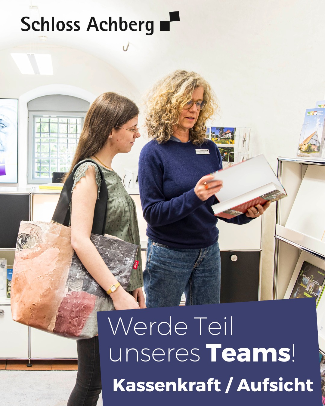 Dieser Alternativtext wurde KI-generiert: Zwei Personen betrachten in einem Innenraum gemeinsam ein offenes Buch oder eine Broschüre. Im Hintergrund befinden sich Poster und eine Anzeige für offene Stellen.