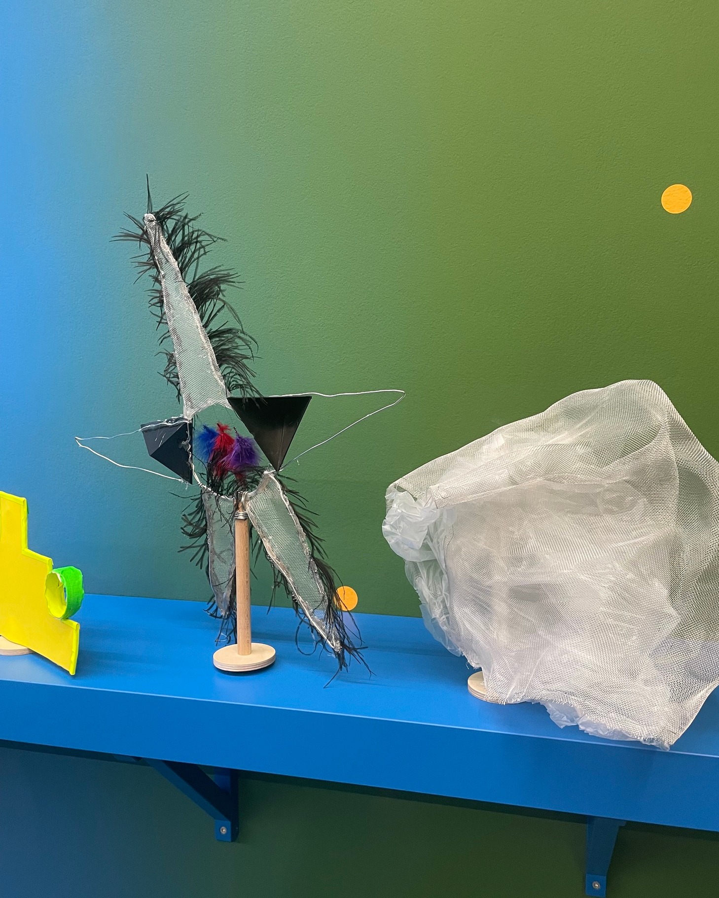 Auf einer blauen Ablage vor einer blau-grün verlaufenden Wand stehen eine gelbe Holzform, eine Figur aus schwarzen Federn, weißem Netz und schwarzen Dreiecken auf einem Holzstab sowie ein weißes Netz. Zwei gelbe Kreise zieren die Wand. (KI-Alternativtext)