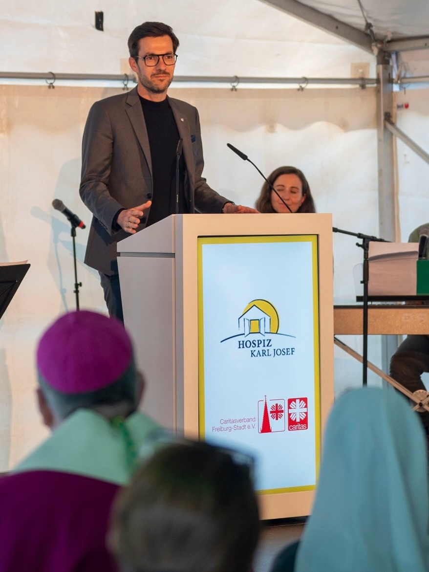 Dieser Alternativtext wurde KI-generiert: Eine Person steht an einem Pult und spricht in ein Mikrofon. Im Hintergrund sind weitere Personen und ein Logo auf dem Pult zu sehen.