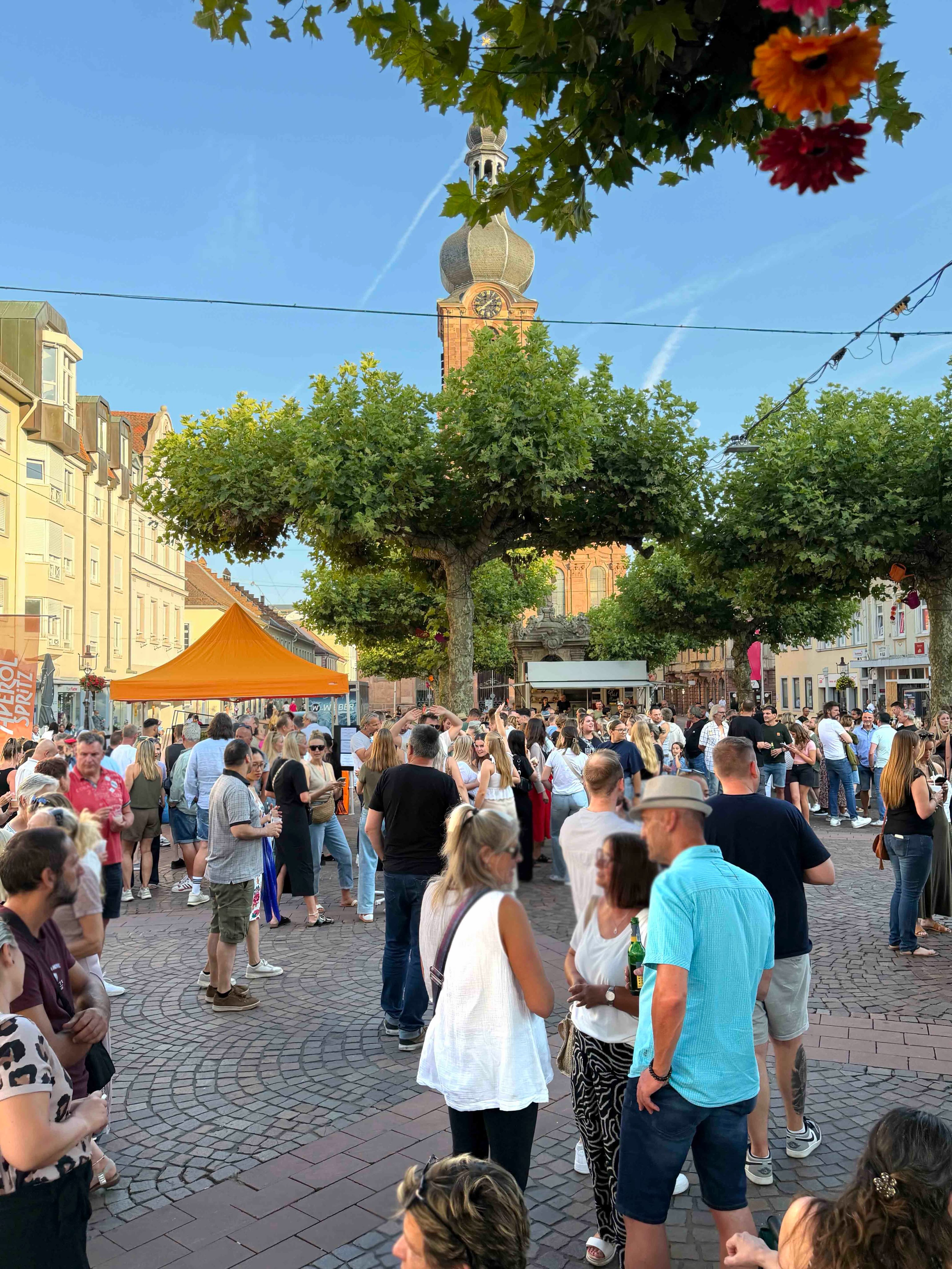 Eine große Menschenmenge versammelt sich auf einem gepflasterten Platz bei einem Straßenfest unter blauem Himmel. Im Hintergrund erhebt sich ein markanter Kirchturm mit Zwiebelhaube und Uhr, flankiert von Bäumen und Gebäuden. Links im Bild steht ein orangefarbenes Zelt, während die Personen im Vordergrund und Mittelgrund stehen, trinken und sich unterhalten. (KI-Alternativtext)