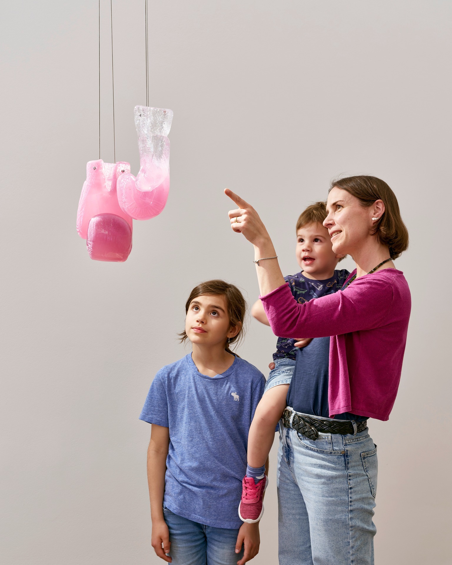 Eine Person hält ein Kind auf dem Arm und zeigt mit dem Finger auf eine hängende Glasinstallation, die wie rosa Boxhandschuhe aussieht. Ein weiteres Kind steht daneben und schaut ebenfalls auf das Kunstwerk. (KI-Alternativtext)