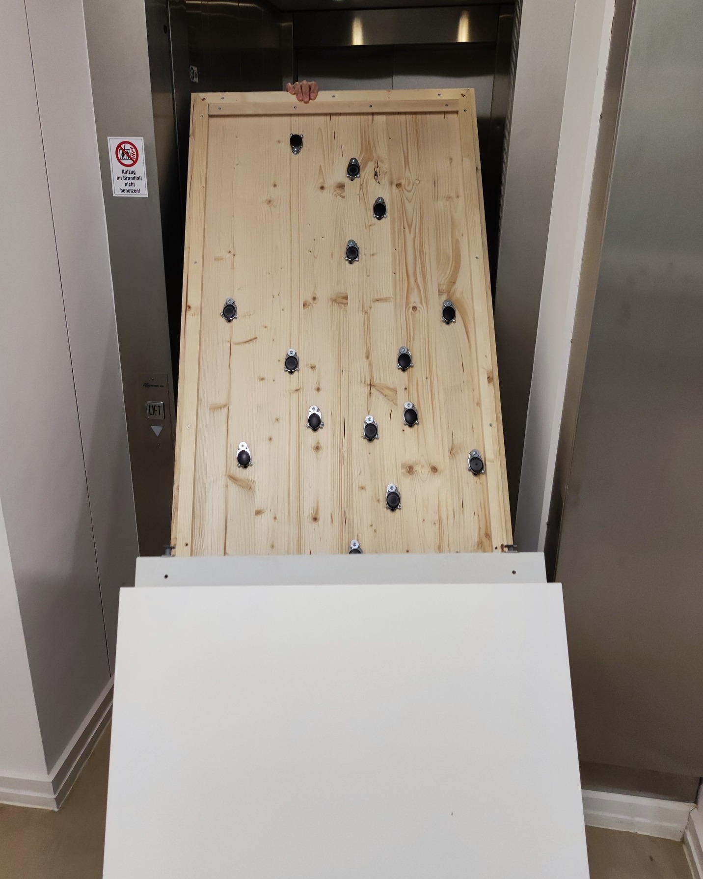 Dieser Alternativtext wurde KI-generiert: Im Inneren eines Aufzugs befindet sich eine Holztür mit mehreren Metallbeschlägen. Die Wände des Aufzugs sind hell.