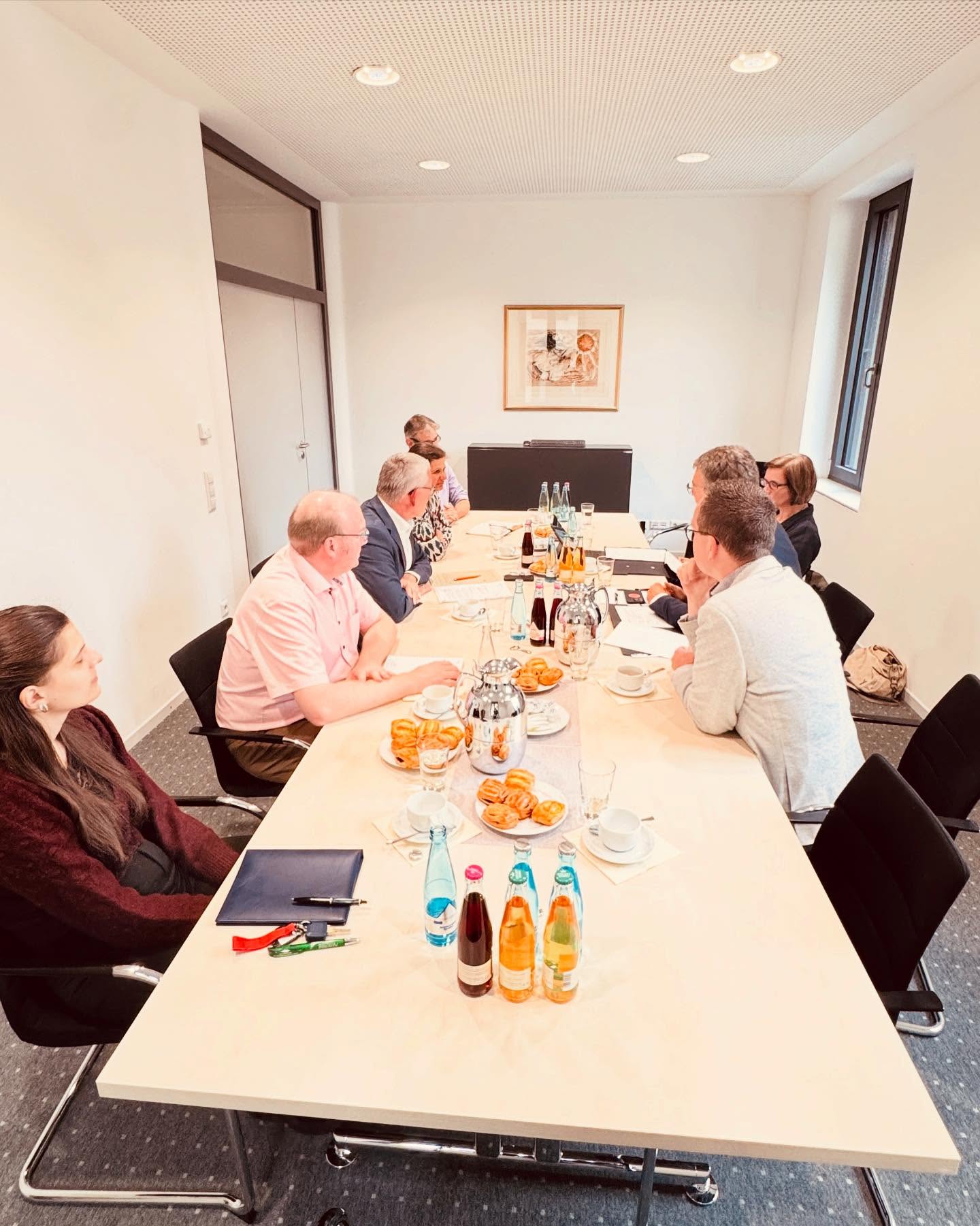 Mehrere Leute sitzen an einem Tisch in einem Konferenzraum. Auf dem Tisch stehen Getränke und Essen bereit.
