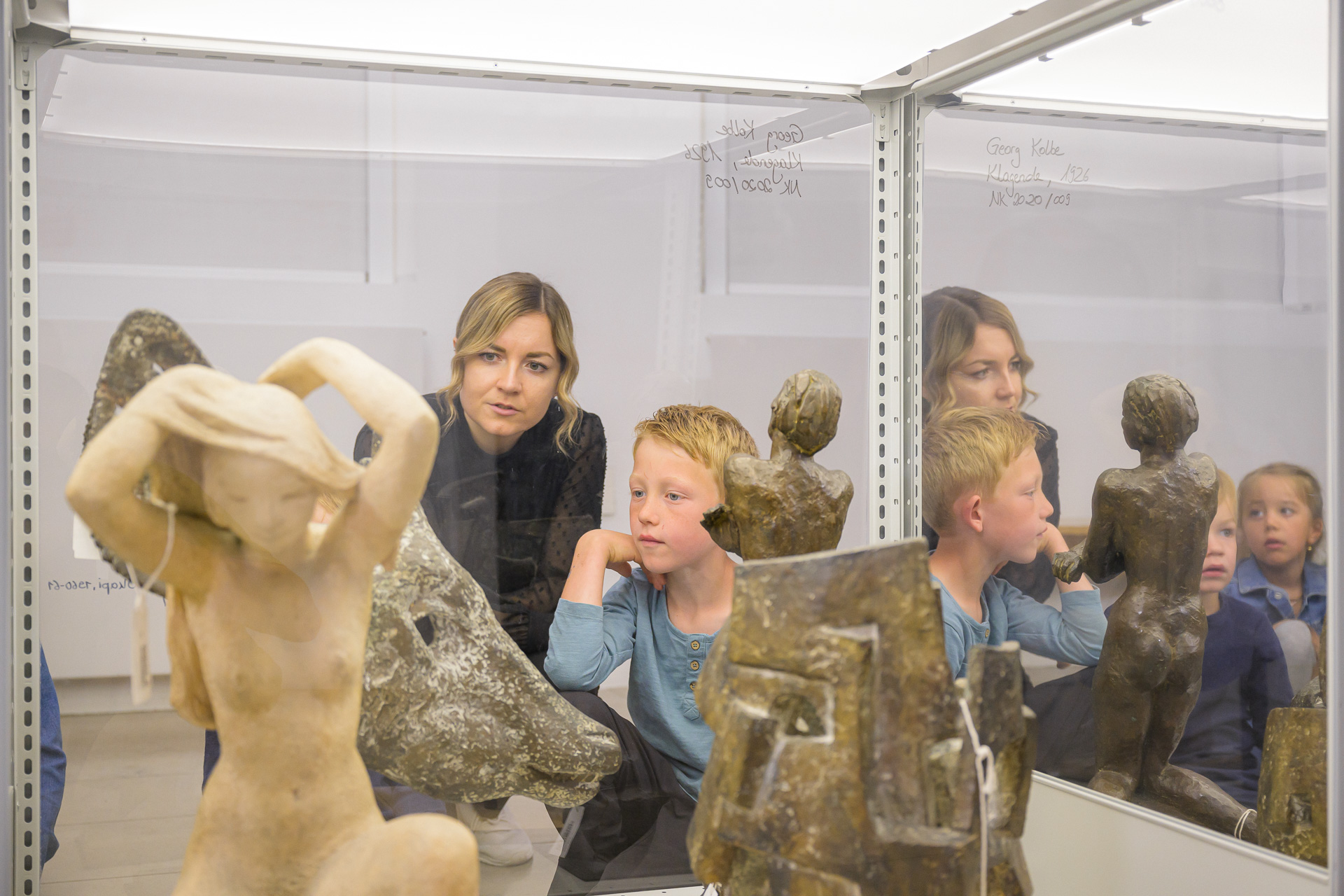 Mehrere Skulpturen, darunter eine helle Figur mit erhobenen Armen und dunklere Figuren, stehen in einer Glasvitrine. Dahinter blicken ein Erwachsener und zwei Kinder durch die Scheibe auf die Kunstwerke. Auf dem Glas steht handschriftlich „Georg Kolbe, Klagenfurt, 1926“. (KI-Alternativtext)