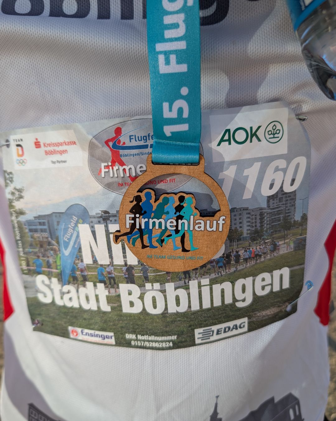 Eine silberne Medaille mit einem blauen Band hängt über einen Startnummer-Aufkleber eines Firmenlaufs in der Stadt Böblingen. Der Aufkleber zeigt die Nummer 1160, das Logo "Firmenlauf im Team gesund und fit" sowie Sponsorenlogos von AOK, Kreissparkasse Böblingen und EDAG. (KI-Alternativtext)