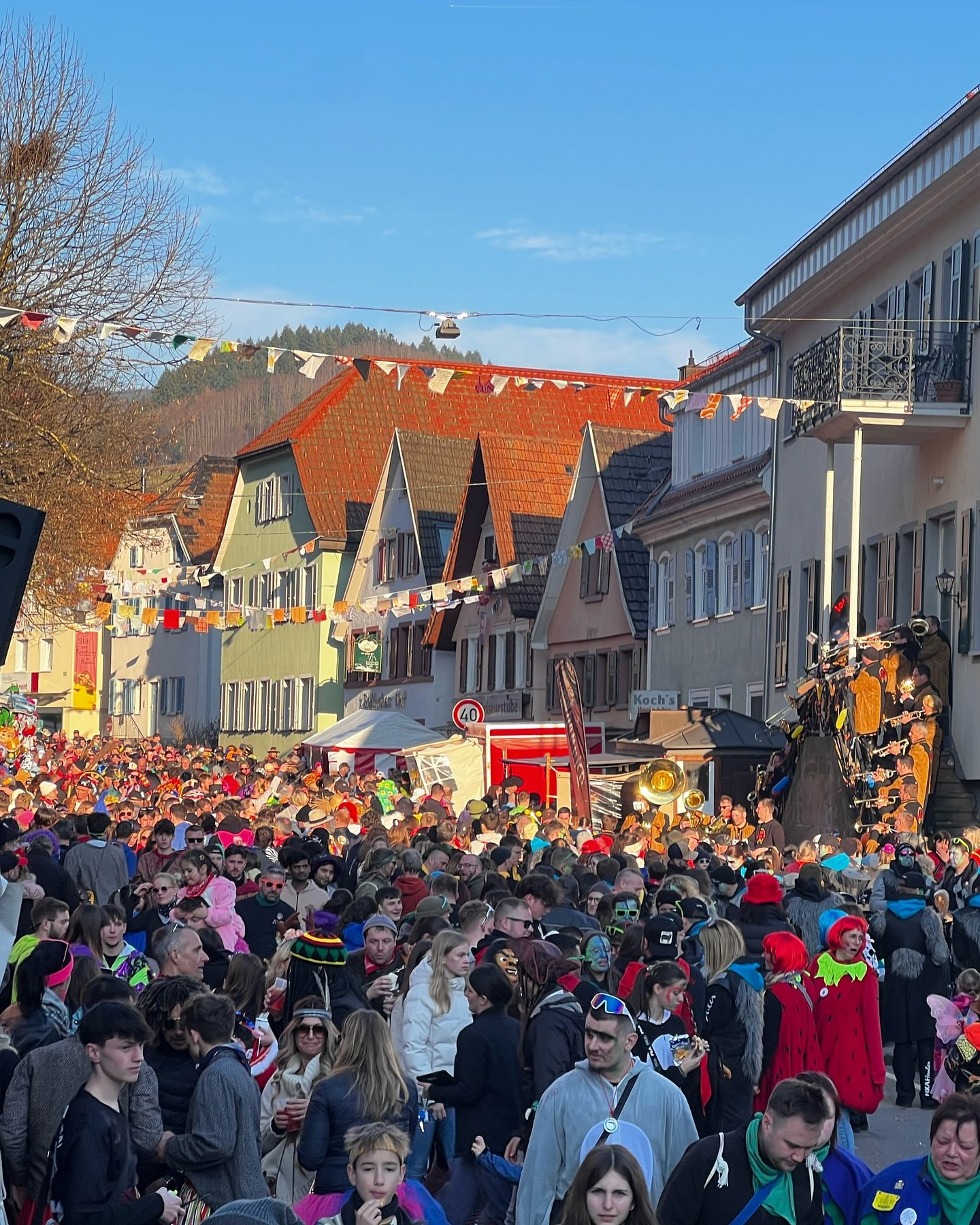 Ein farbenfrohes Bild zeigt eine große Menschenmenge auf einer Straße, gesäumt von Gebäuden. Dekorative Elemente sind über der Straße angebracht.