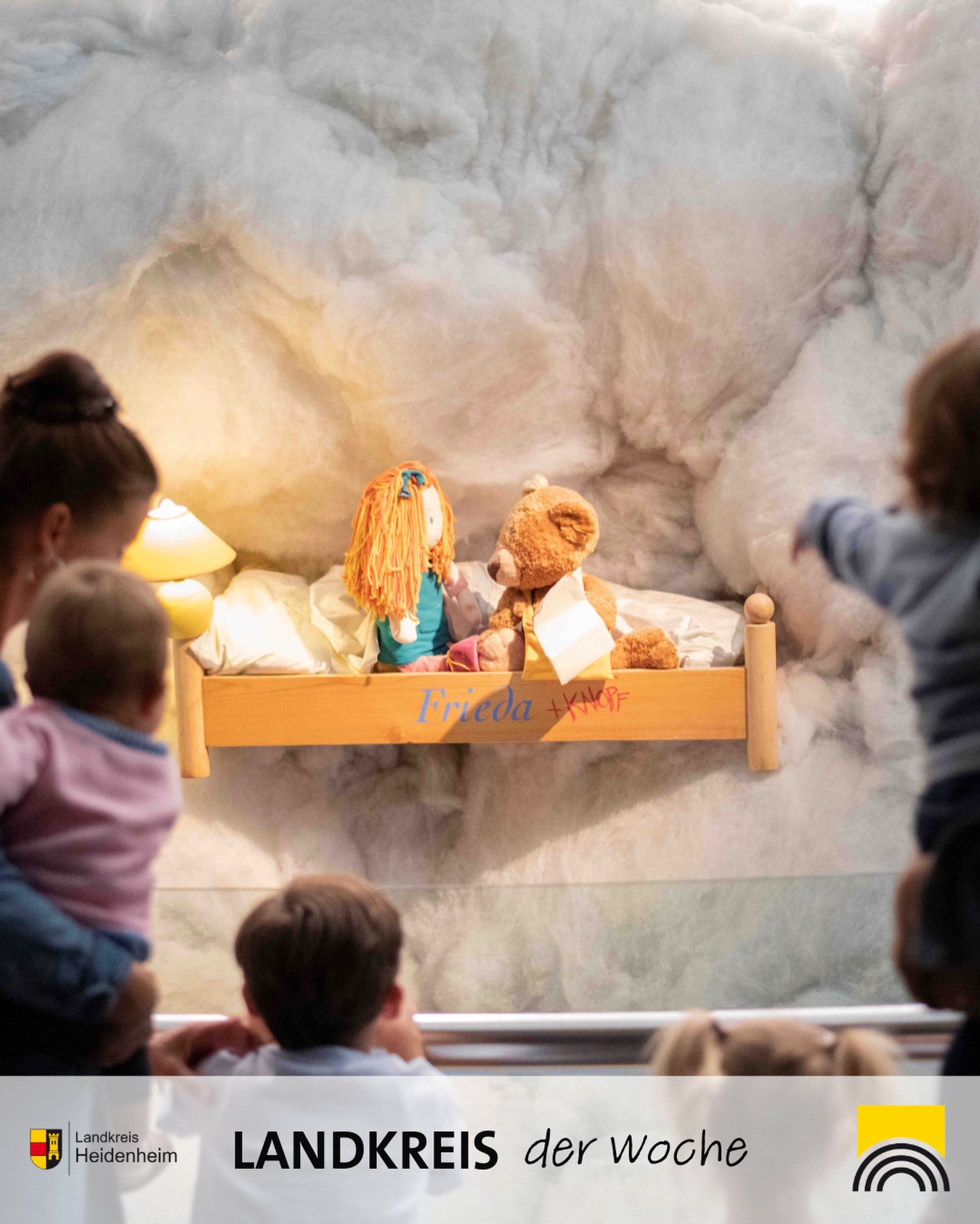 Dieser Alternativtext wurde KI-generiert: Ein hölzernes Regalbrett mit Stofftieren und einer Puppe ist an einer Wand befestigt. Im Hintergrund sind Teile von Köpfen und Schultern zu sehen.