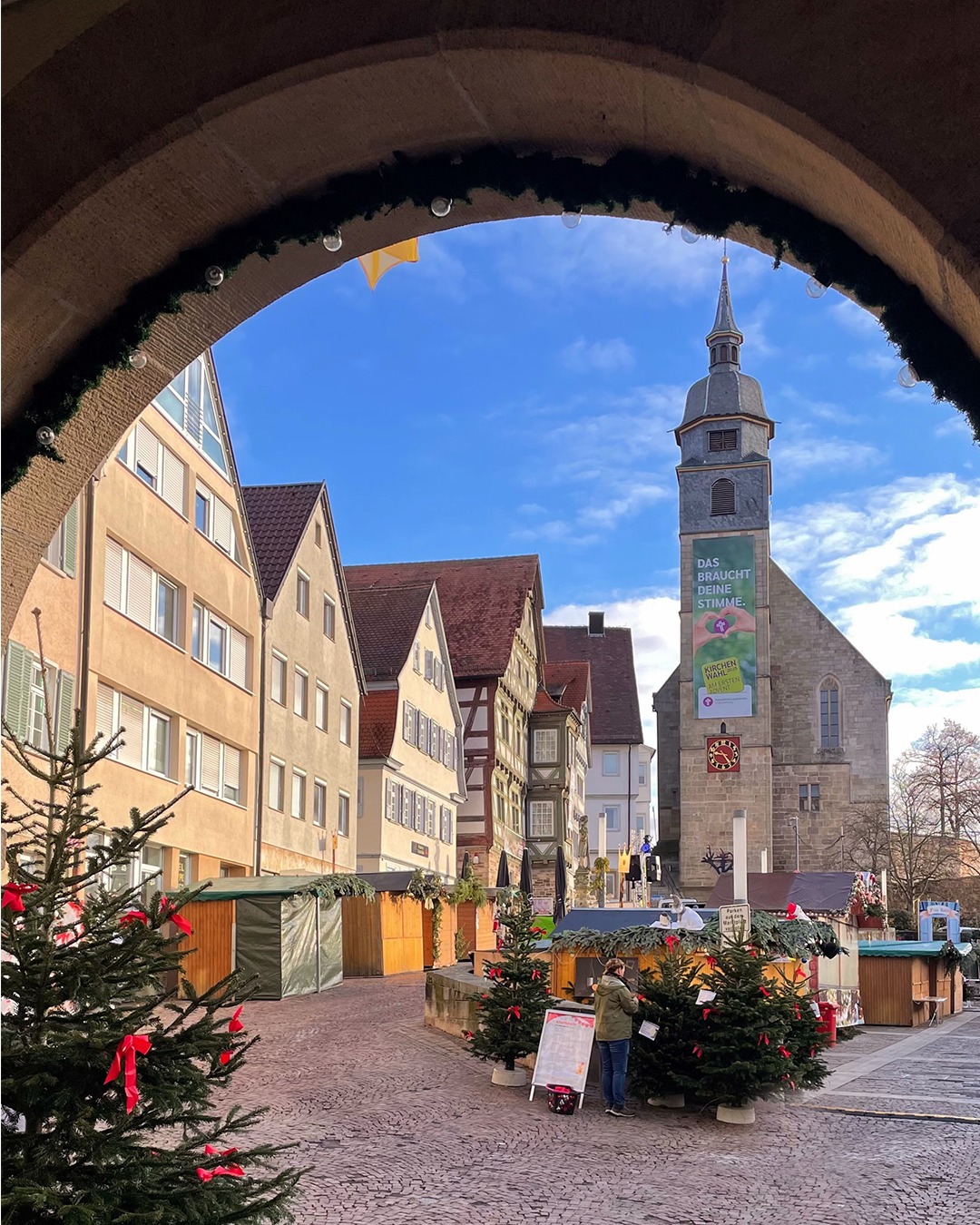 Dieser Alternativtext wurde KI-generiert: Ein Blick durch einen runden Rahmen auf eine Stadtansicht mit Fachwerkhäusern und einer Kirche. Im Vordergrund befinden sich weihnachtlich dekorierte Bäume und Marktstände.