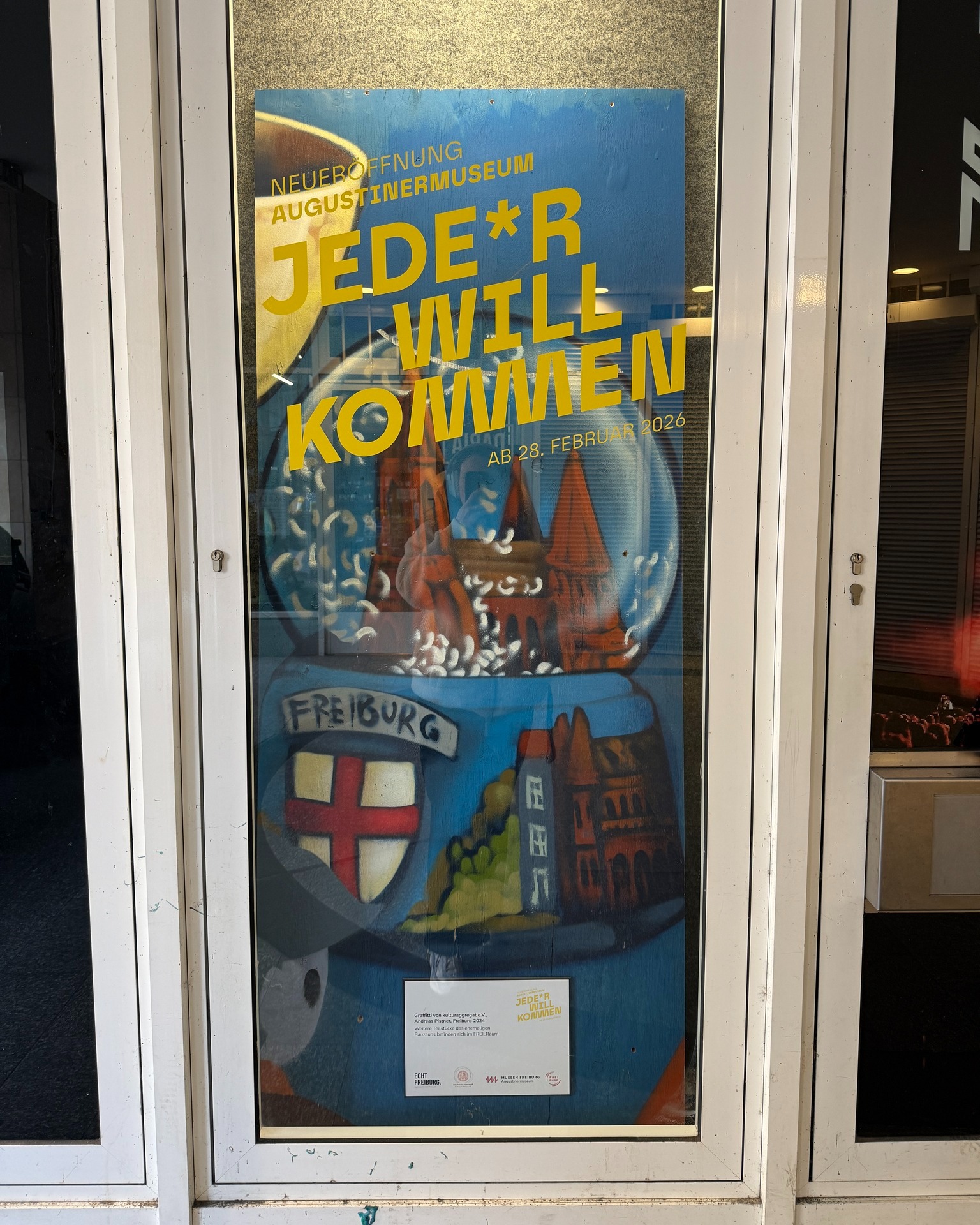 Dieser Alternativtext wurde KI-generiert: Ein farbenfrohes Plakat vor einem Glastür, das eine Ankündigung mit dem Datum 28. Februar 2026 enthält. Das Plakat zeigt stilisierte Gebäude und Elemente, die auf eine Stadt hinweisen.