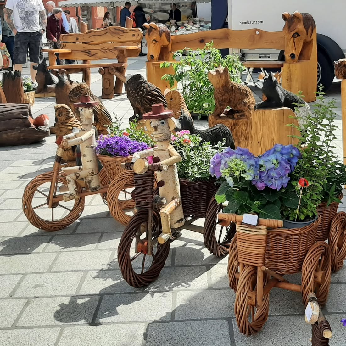 Bild von handgeschnitzten Holzfiguren und Blumenkübeln auf einem Markt. Die Figuren sitzen auf kleinen, geflochtenen Dreirädern.