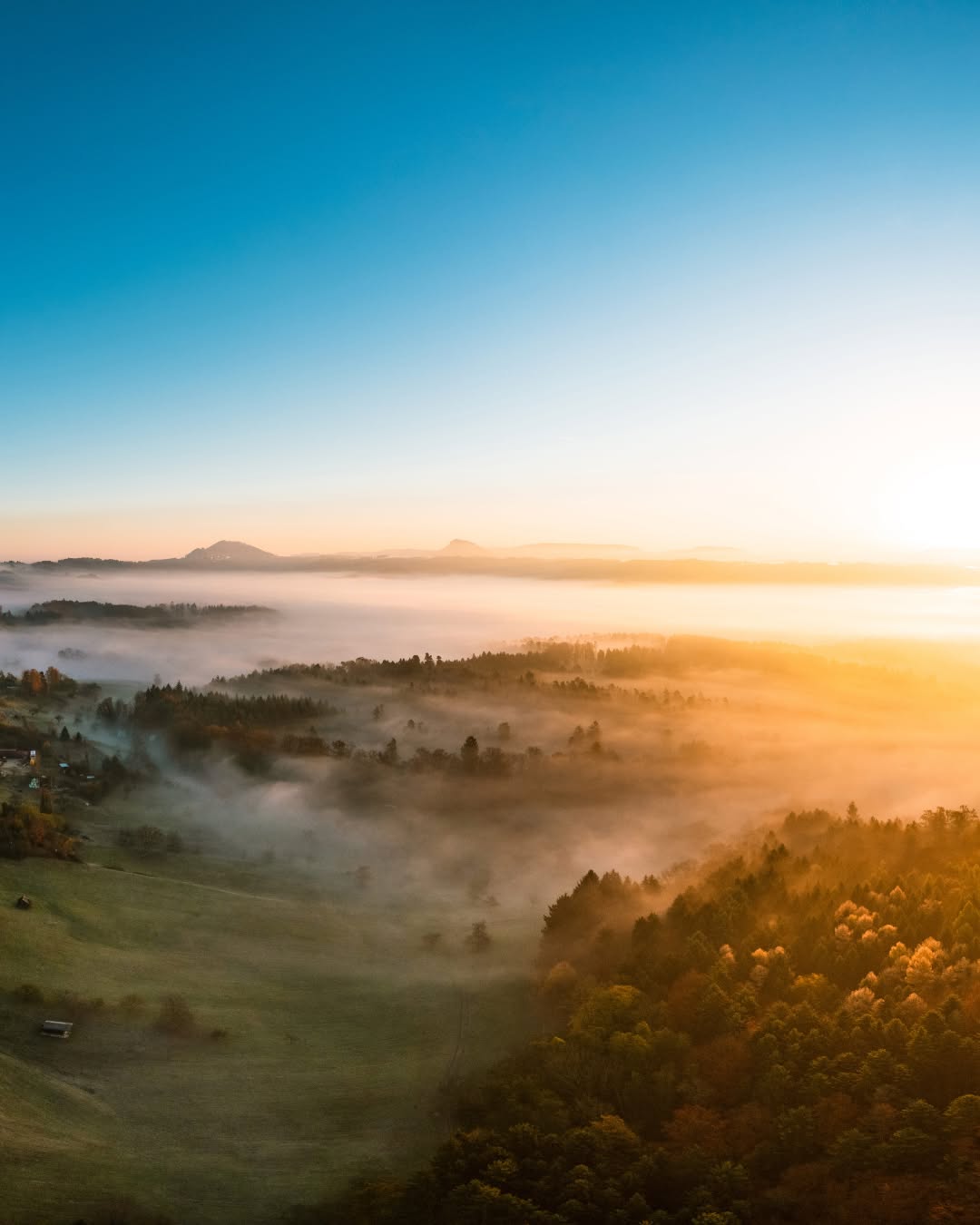 Dieser Alternativtext wurde KI-generiert: Eine Landschaft mit Wiesen und Wäldern liegt in einem Nebel. Die Sonne geht auf und taucht die Szene in warmes Licht.