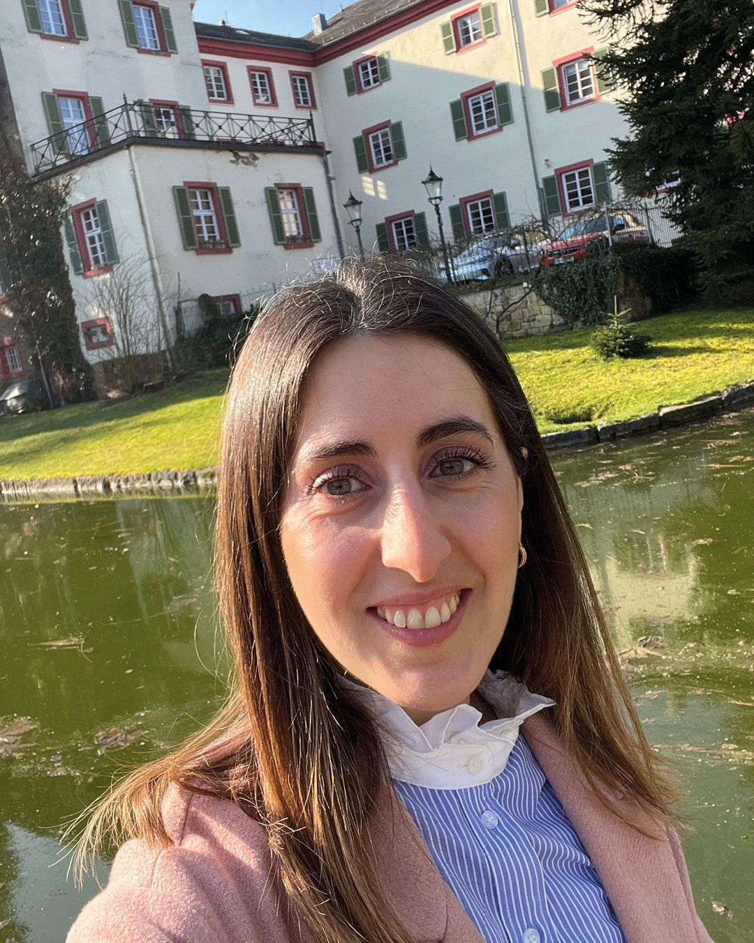 Dieser Alternativtext wurde KI-generiert: Eine Person macht ein Selfie vor einem Gebäude mit vielen Fenstern und grünem Bewuchs. Im Vordergrund befindet sich ein Teich, der das Gebäude und die Person widerspiegelt.