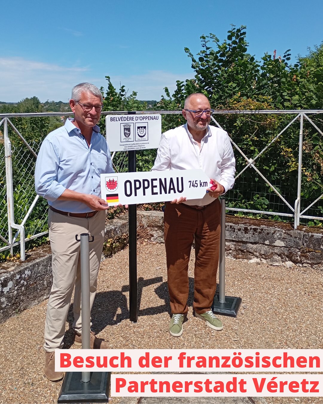 Zwei Personen stehen neben einem Schild mit der Aufschrift "Oppenau 745 km". Im Hintergrund sind Bäume und ein blauer Himmel zu sehen.