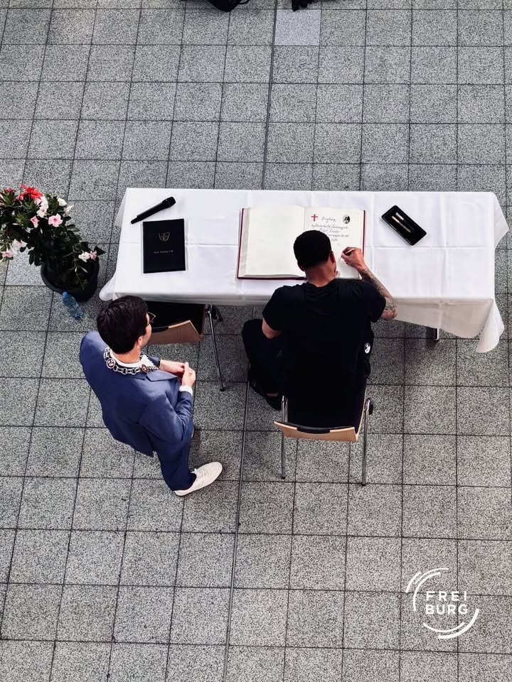 Zwei Personen sitzen an einem mit weißer Tischdecke bedeckten Tisch auf gepflastertem Boden. Eine Person unterschreibt ein offenes Buch mit rotem Einband, daneben liegt ein schwarzes Buch und ein Stift. Die andere Person, mit Amtskette, beobachtet das Geschehen. Eine Blumenvase steht links neben dem Tisch. (KI-Alternativtext)