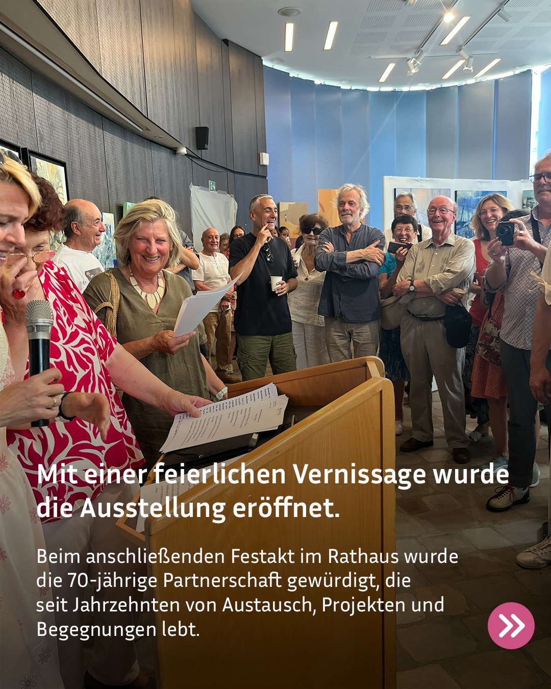 Eine Person spricht an einem hölzernen Rednerpult in ein Mikrofon, während eine weitere Person daneben steht und Papiere hält. Im Hintergrund steht eine Gruppe von Menschen in einem Ausstellungsraum, die zuhören und Fotos machen. Der Text im Bild lautet: „Mit einer feierlichen Vernissage wurde die Ausstellung eröffnet. Beim anschließenden Festakt im Rathaus wurde die 70-jährige Partnerschaft gewürdigt, die seit Jahrzehnten von Austausch, Projekten und Begegnungen lebt.“ (KI-Alternativtext)