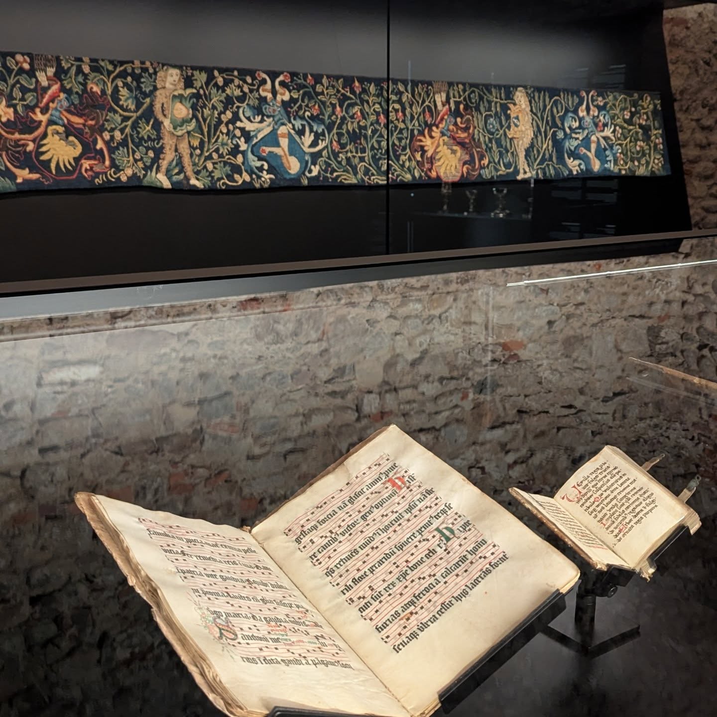 Dieser Alternativtext wurde KI-generiert: Ein geöffnetes Buch mit mittelalterlicher Schrift und kunstvolle Wandteppiche sind in einem Raum mit Steinwänden zu sehen. Das Bild ist farbig und zeigt historische Gegenstände in einer Museumsumgebung.