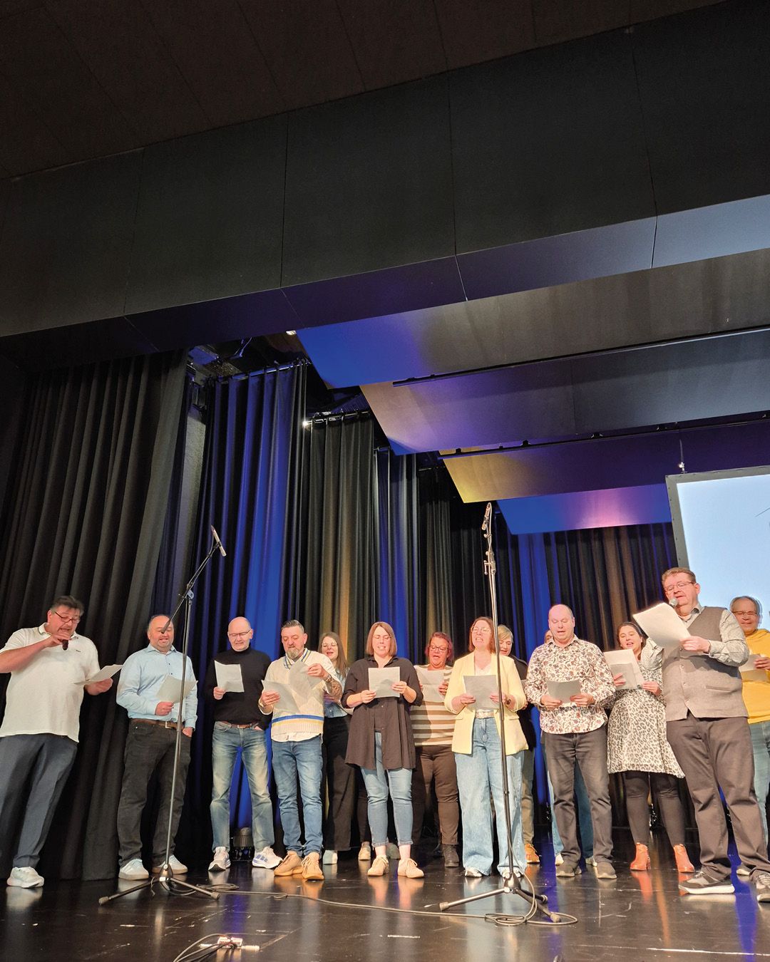 Dieser Alternativtext wurde KI-generiert: Eine Gruppe von Personen steht auf einer Bühne vor einem Vorhang mit Mikrofonen und liest von Blättern ab. Die Szene ist in einem Innenraum mit blauer Beleuchtung aufgenommen.