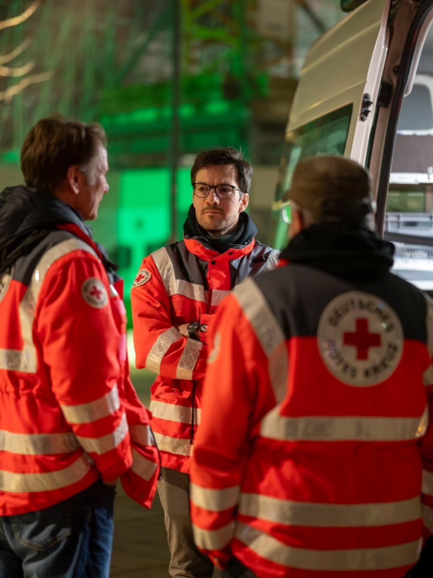 Dieser Alternativtext wurde KI-generiert: Mehrere Personen mit Warnwesten stehen im Freien zusammen. Im Hintergrund ist ein Fahrzeug und grüne Beleuchtung zu sehen.