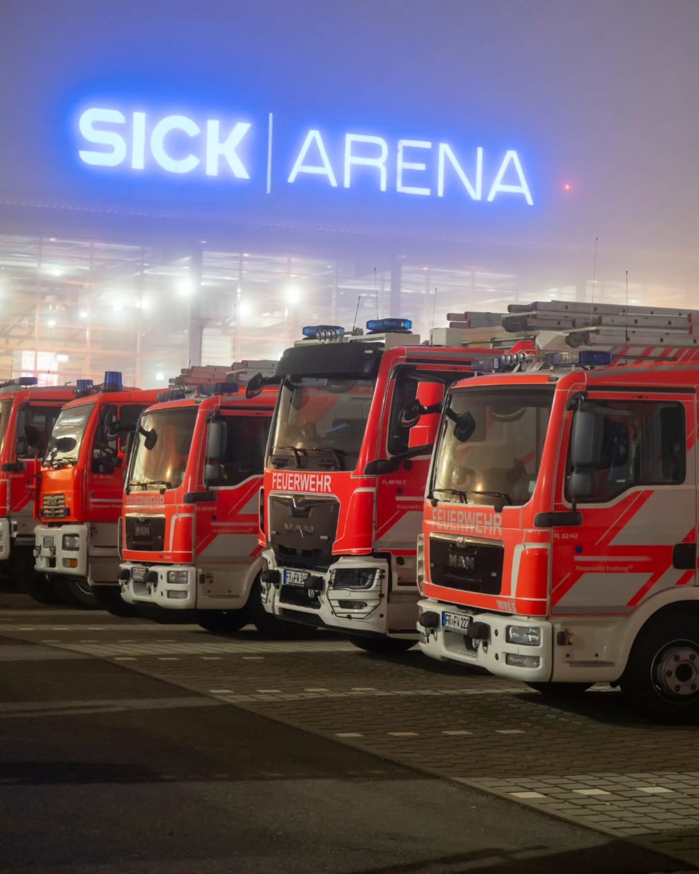 Dieser Alternativtext wurde KI-generiert: Mehrere rote Einsatzfahrzeuge stehen in einer Reihe vor einem Gebäude mit beleuchtetem Schriftzug. Das Bild ist in der Nacht aufgenommen und zeigt Blaulicht.