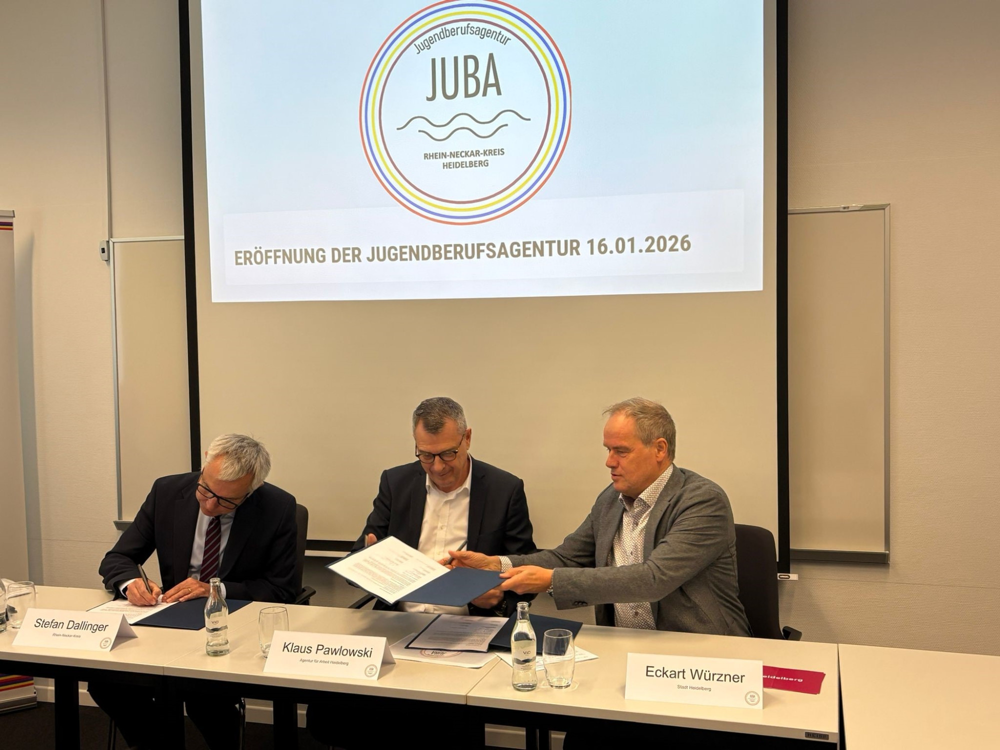 Dieser Alternativtext wurde KI-generiert: Ein farbiges Foto zeigt eine Gruppe von Personen, die an einem Tisch sitzen. Im Hintergrund befindet sich ein Banner mit Text.