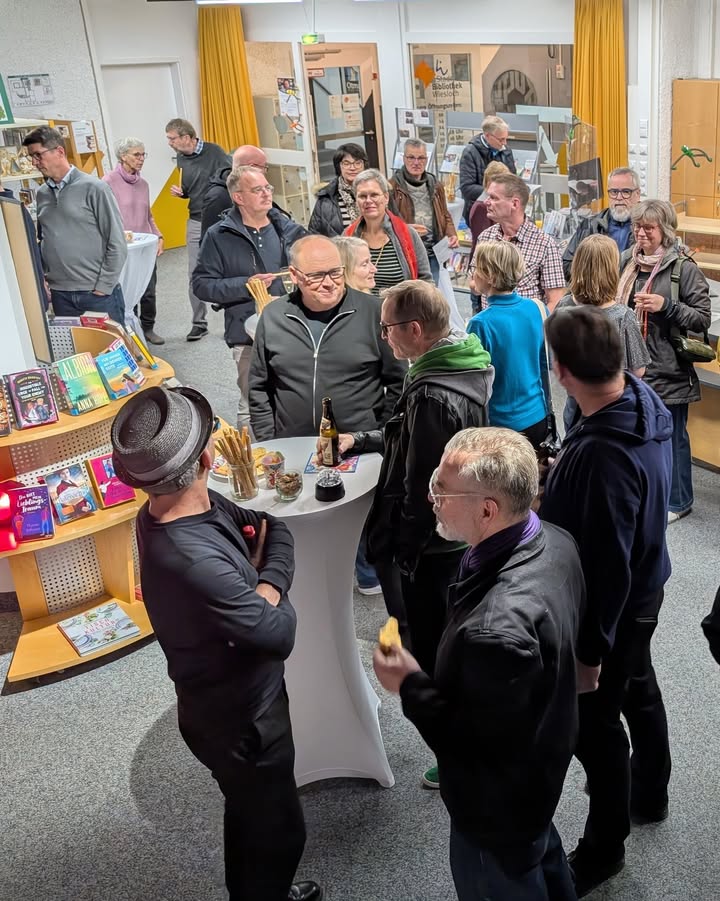Dieser Alternativtext wurde KI-generiert: Eine Gruppe von Menschen steht in einem Raum mit Büchern und Getränken zusammen. Es scheint sich um eine Veranstaltung zu handeln, möglicherweise eine Buchpräsentation.