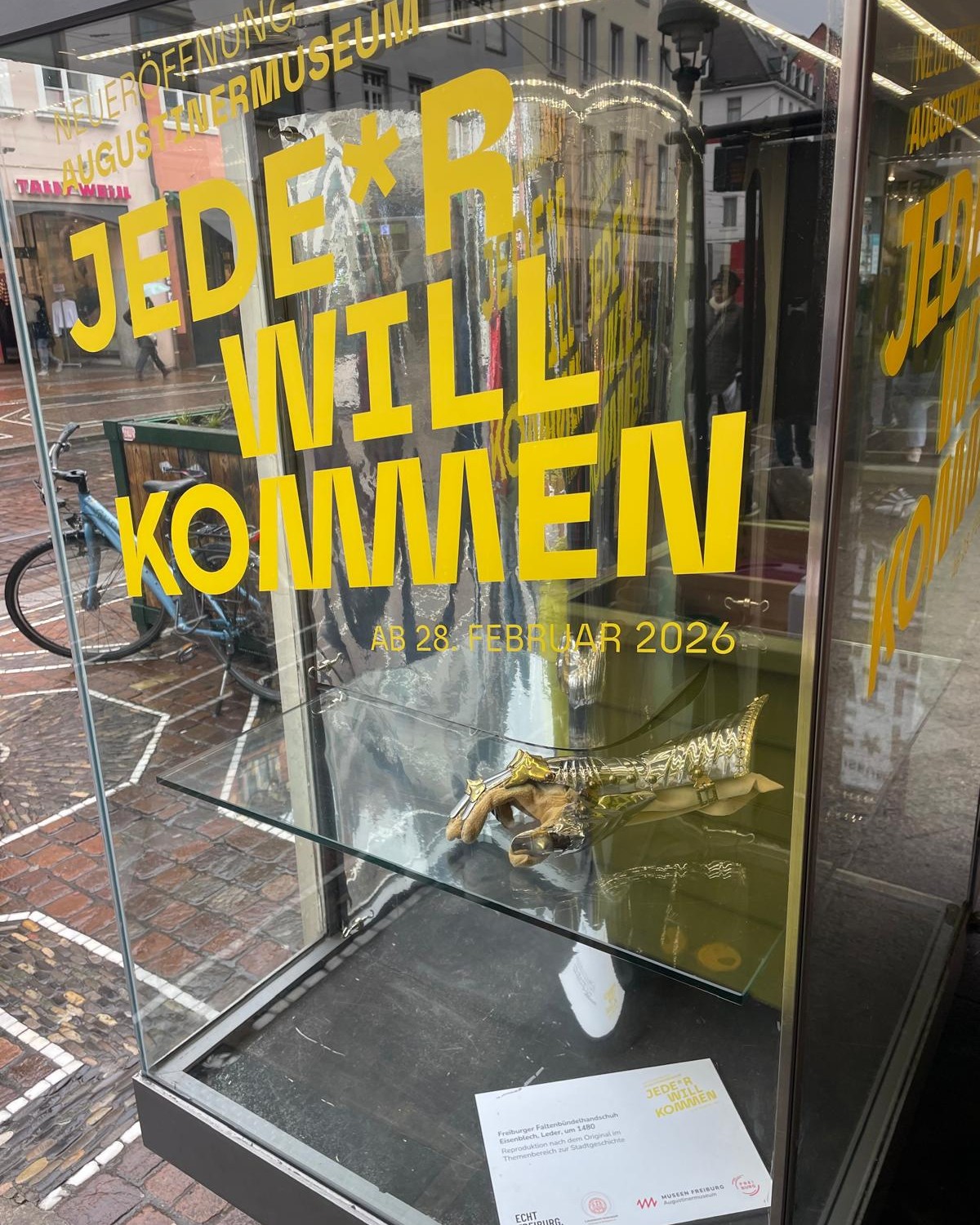 Dieser Alternativtext wurde KI-generiert: Eine Straße mit einem Schaufenster, das Plakate und eine Vogel-Skulptur in einem Glaskasten zeigt. Das Foto ist farbig, die Plakate sind in Gelb und Schwarz gehalten.