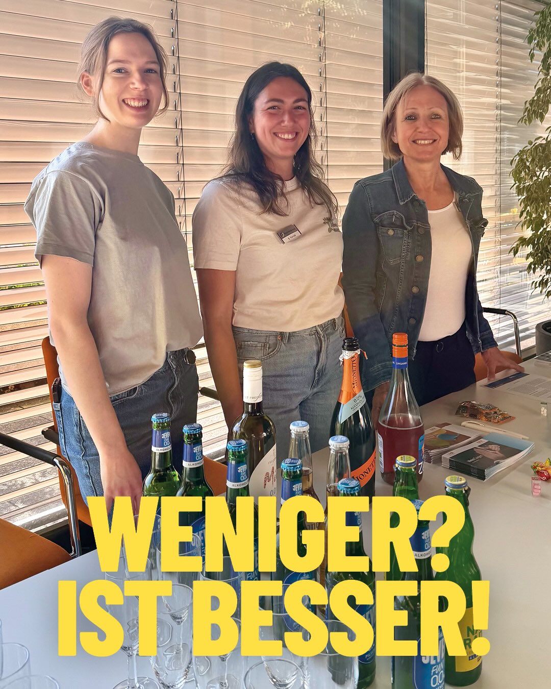 Drei lächelnde Personen stehen hinter einem Tisch, der mit verschiedenen Flaschen und Gläsern gedeckt ist. Im Vordergrund überlagert der große gelbe Schriftzug „WENIGER? IST BESSER!“ das Bild. (KI-Alternativtext)