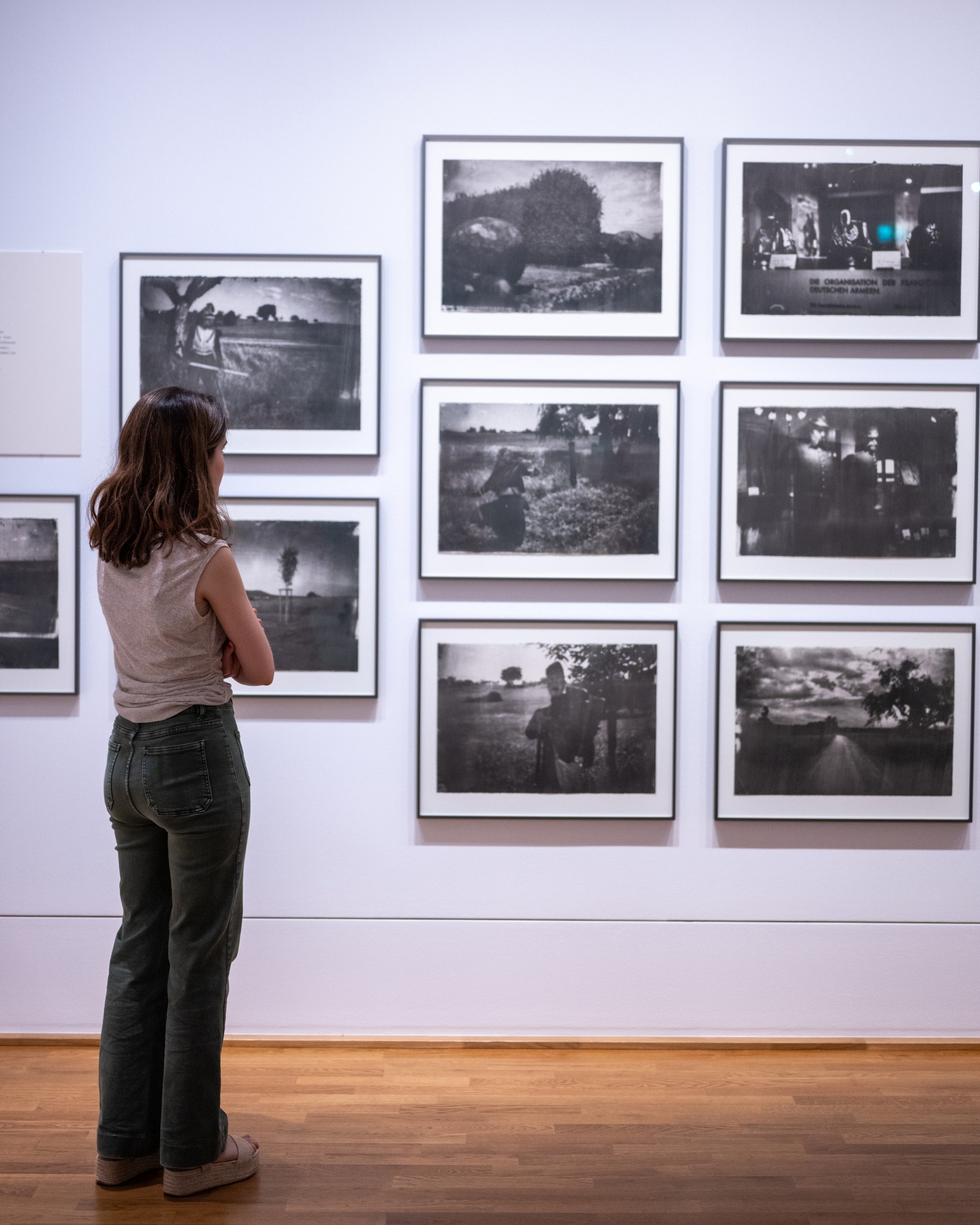 Eine Person mit gekreuzten Armen steht mit dem Rücken zur Kamera und betrachtet eine Wand mit mehreren gerahmten Schwarz-Weiß-Fotografien. Die Person trägt ein ärmelloses Oberteil und eine Jeans. Auf dem Foto oben rechts ist der Text „DIE ORGANISATION DER FRANZÖSISCHEN DEUTSCHEN ARMEE“ lesbar. (KI-Alternativtext)