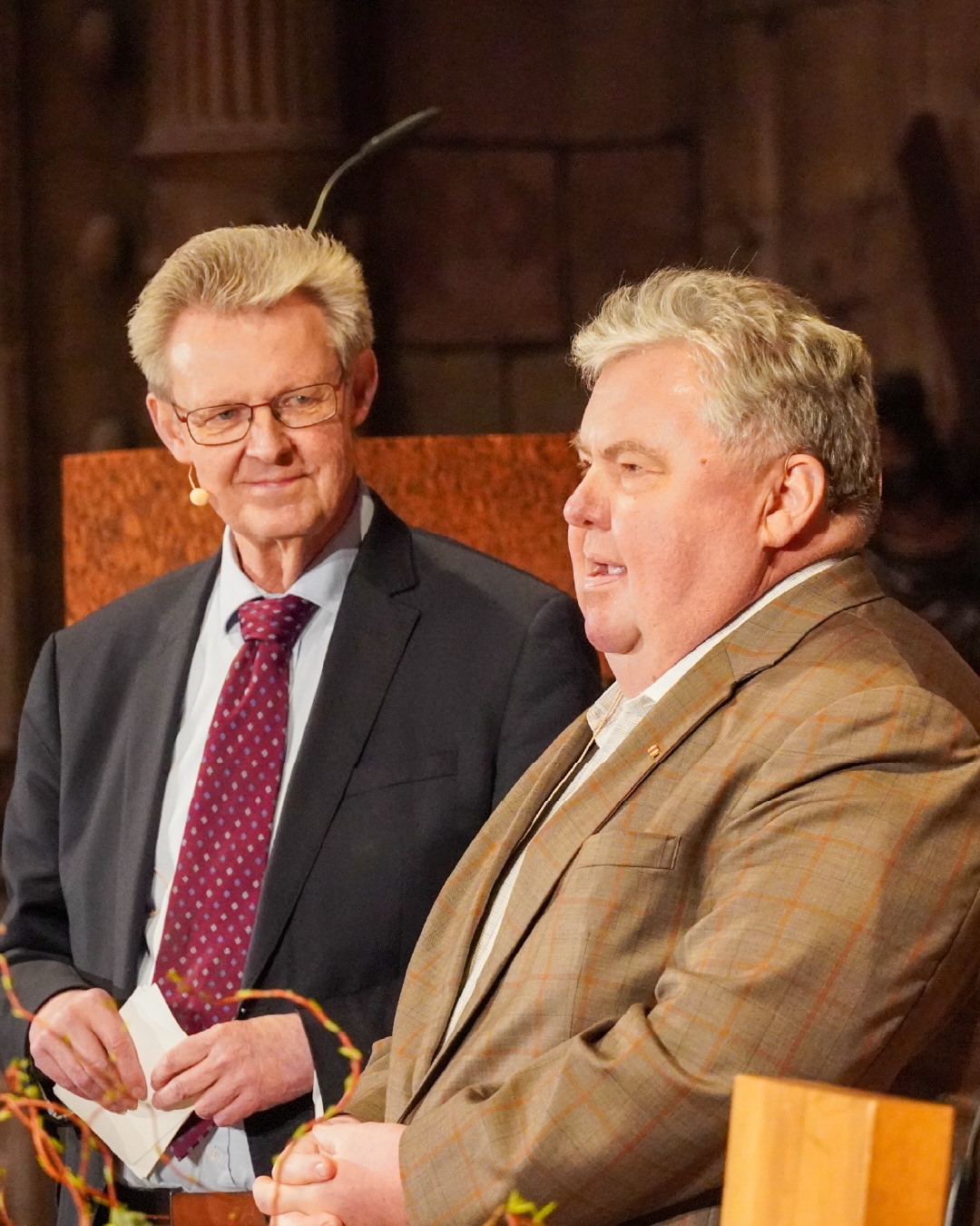 Dieser Alternativtext wurde KI-generiert: Zwei Personen mit Anzug und Krawatte sitzen nebeneinander und sprechen. Im Hintergrund ist eine Wand aus Stein zu sehen.