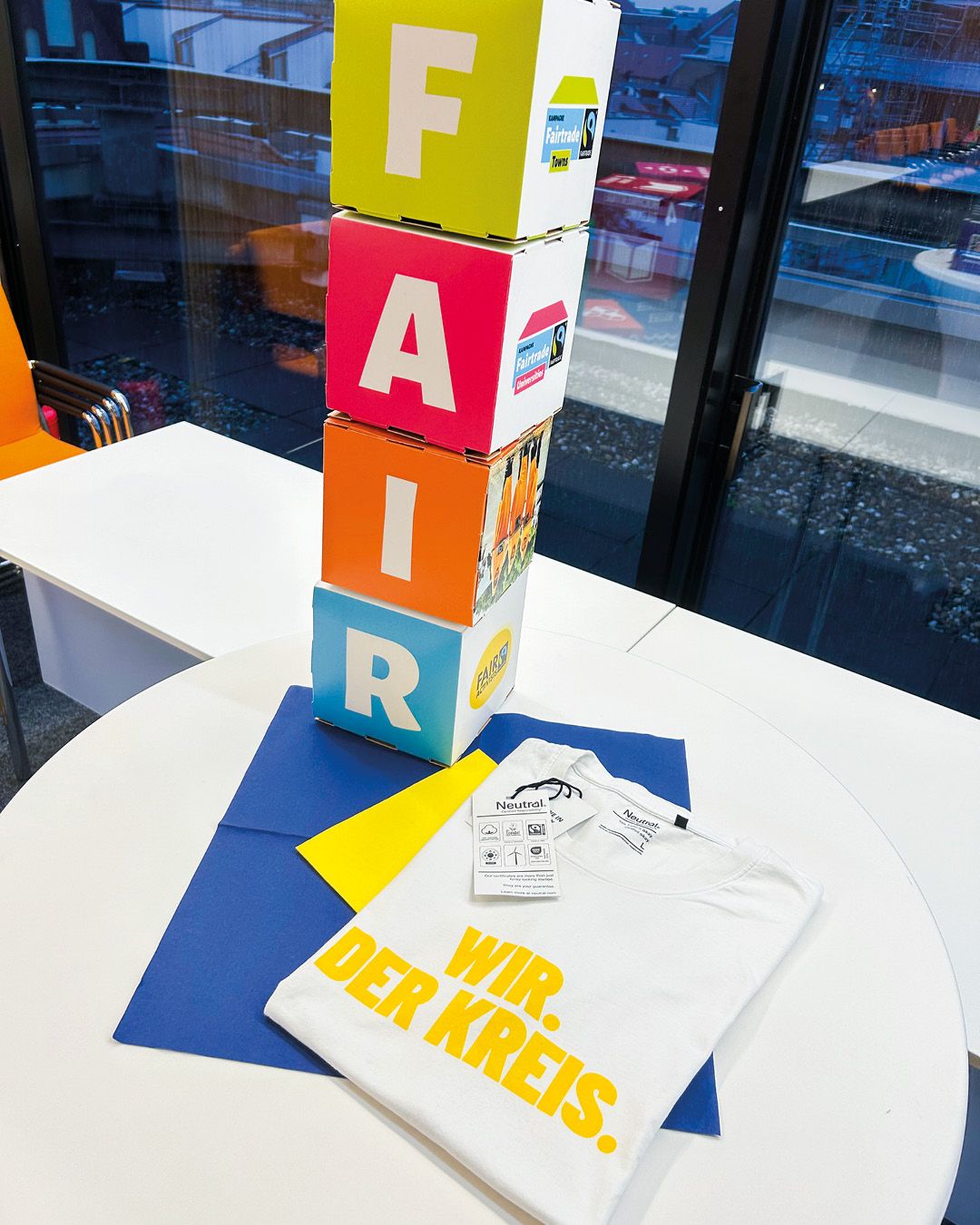 Dieser Alternativtext wurde KI-generiert: Ein Stapel farbiger Kartonwürfel mit Buchstaben steht auf einem runden Tisch. Daneben liegen ein weißes T-Shirt und ein blaues Blatt Papier mit Aufdruck.