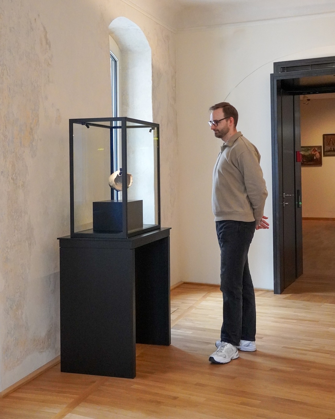 Dieser Alternativtext wurde KI-generiert: Ein Innenraum mit einer Person, die sich einen Ausstellungsgegenstand in einer Vitrine ansieht. Der Raum ist hell erleuchtet und verfügt über einen Holzboden.