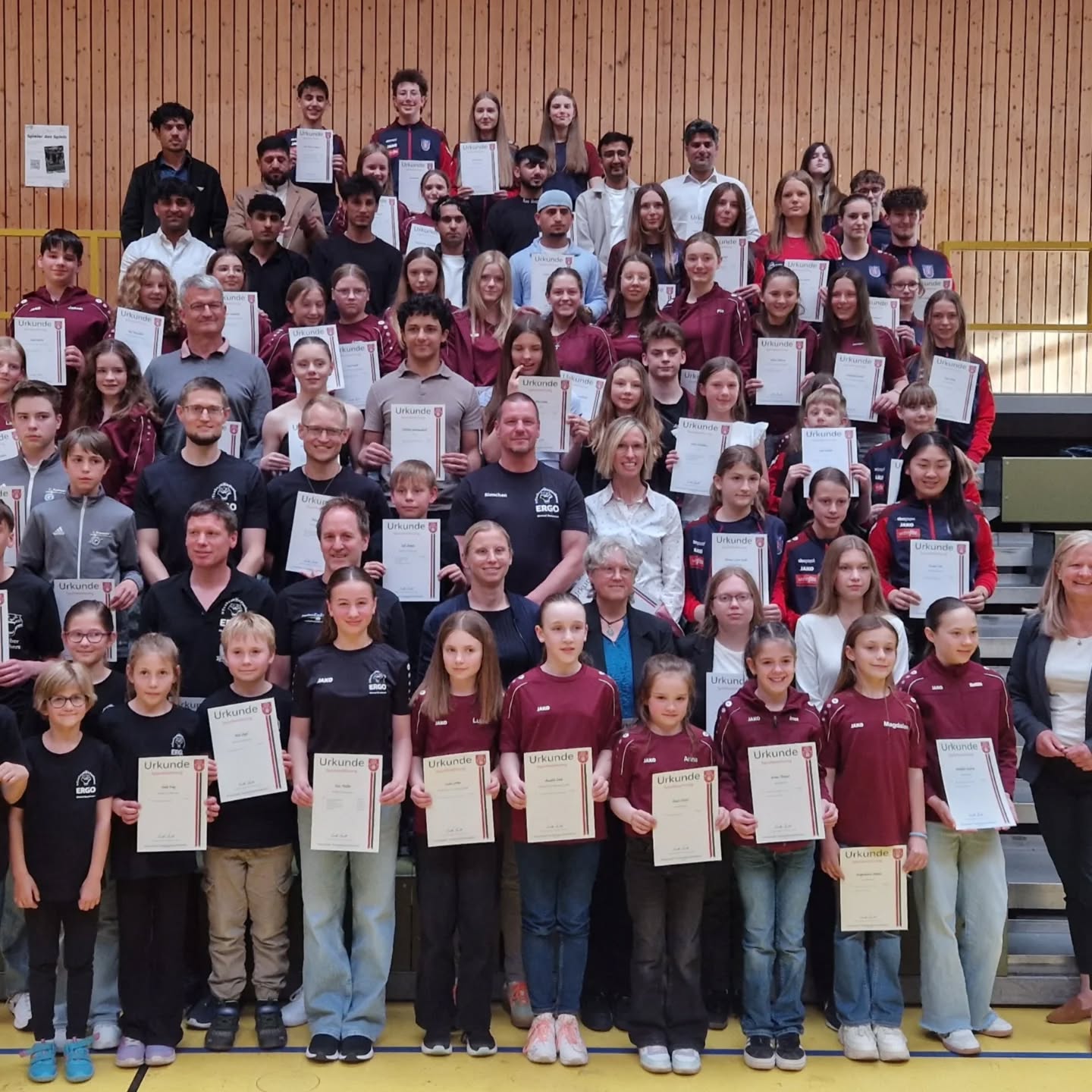 Dieser Alternativtext wurde KI-generiert: Eine große Gruppe von Personen posiert für ein Foto und hält Urkunden in den Händen. Die Aufnahme ist farbig und zeigt die Menschen in einer Sporthalle.