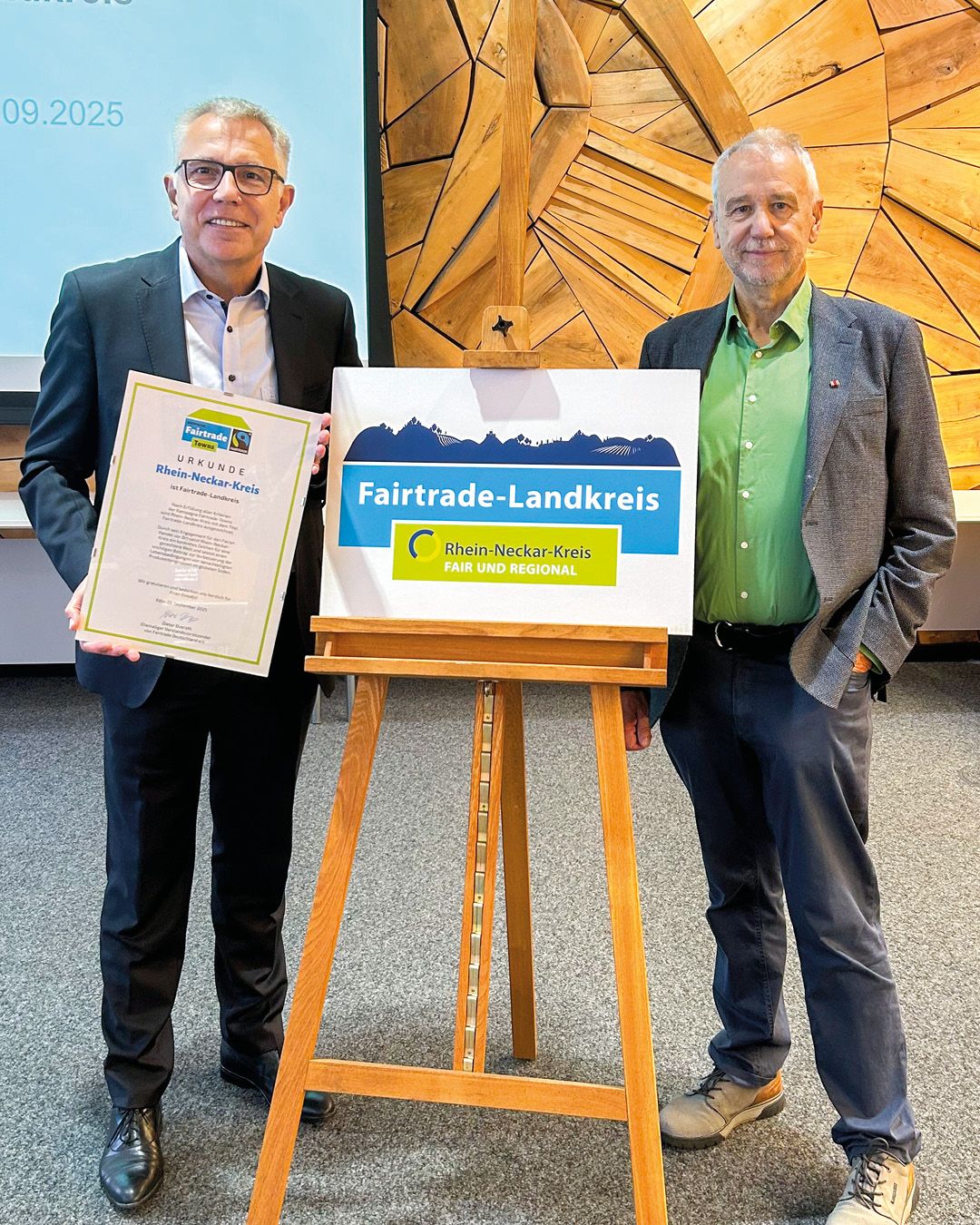 Dieser Alternativtext wurde KI-generiert: Zwei Personen stehen vor einem Holzständer mit einem Schild, auf dem ein Bergmotiv und ein Fairtrade-Logo zu sehen sind. Im Hintergrund befindet sich eine helle Wand mit einem Teppichmuster.