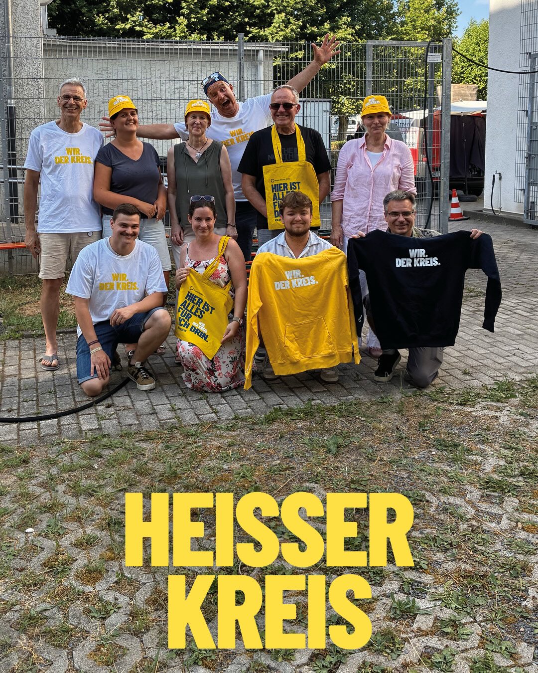Eine Gruppe von Menschen posiert im Freien und hält Kleidungsstücke sowie Taschen hoch, auf denen „WIR. DER KREIS.“ und „HIER IST ALLES FÜR DICH DRIN.“ steht. Im Vordergrund ist groß der gelbe Schriftzug „HEISSER KREIS“ eingeblendet. (KI-Alternativtext)