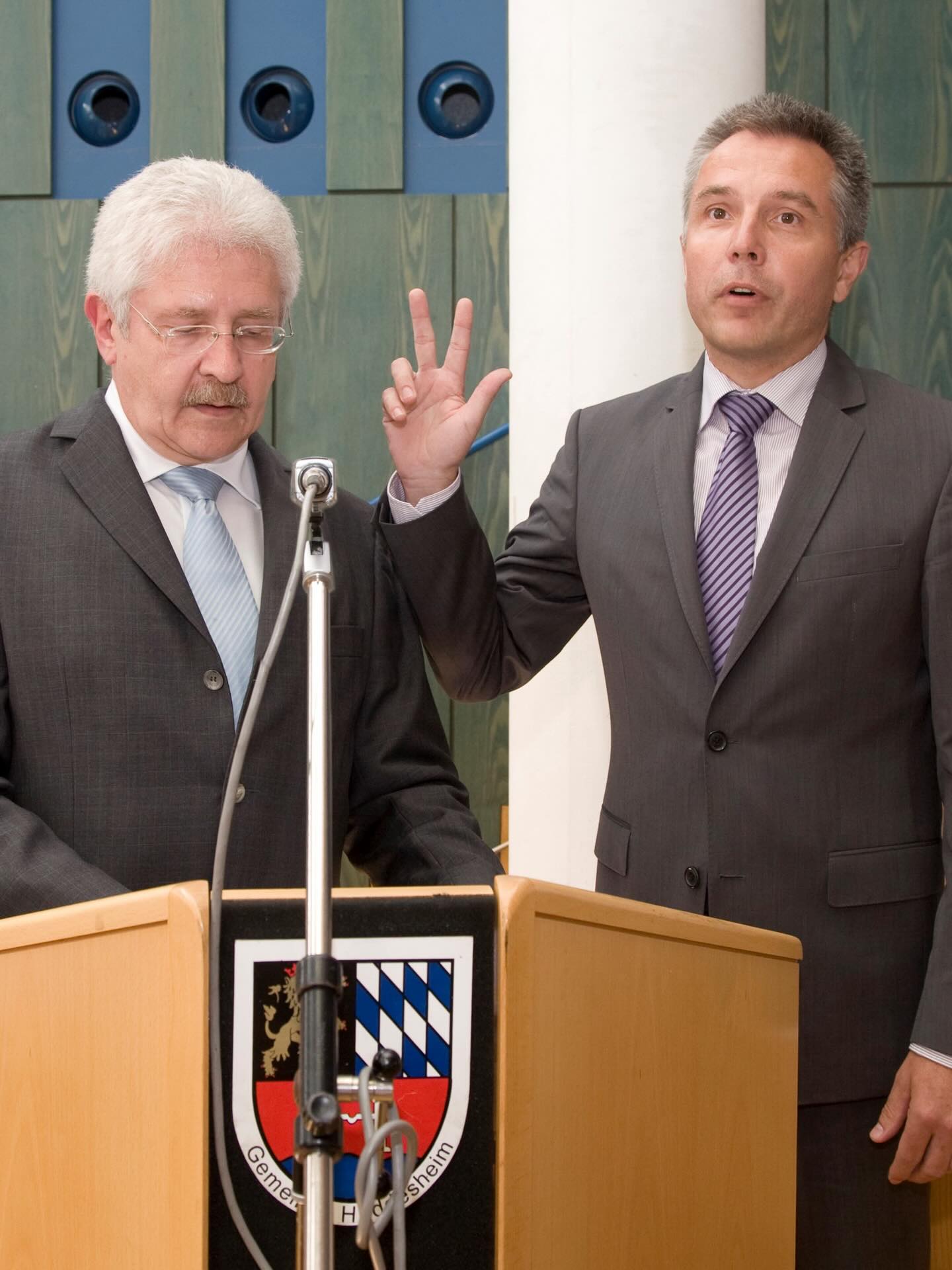 Dieser Alternativtext wurde KI-generiert: Zwei Personen in Anzügen stehen hinter einem Pult mit einem Wappen. Eine Person hebt die Hand, während die andere zuhört.