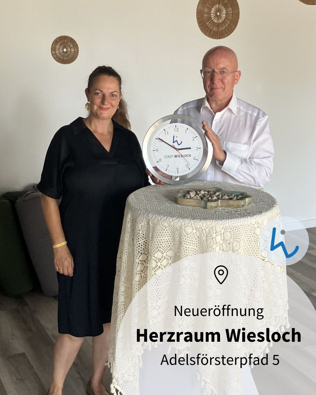 Zwei Personen stehen neben einem Tisch mit weißer Tischdecke, wobei die Person rechts eine große runde Uhr mit der Aufschrift "STADT WIESLOCH" hält. Ein Text-Overlay unten rechts kündigt die "Neueröffnung Herzraum Wiesloch Adelsförsterpfad 5" an. (KI-Alternativtext)