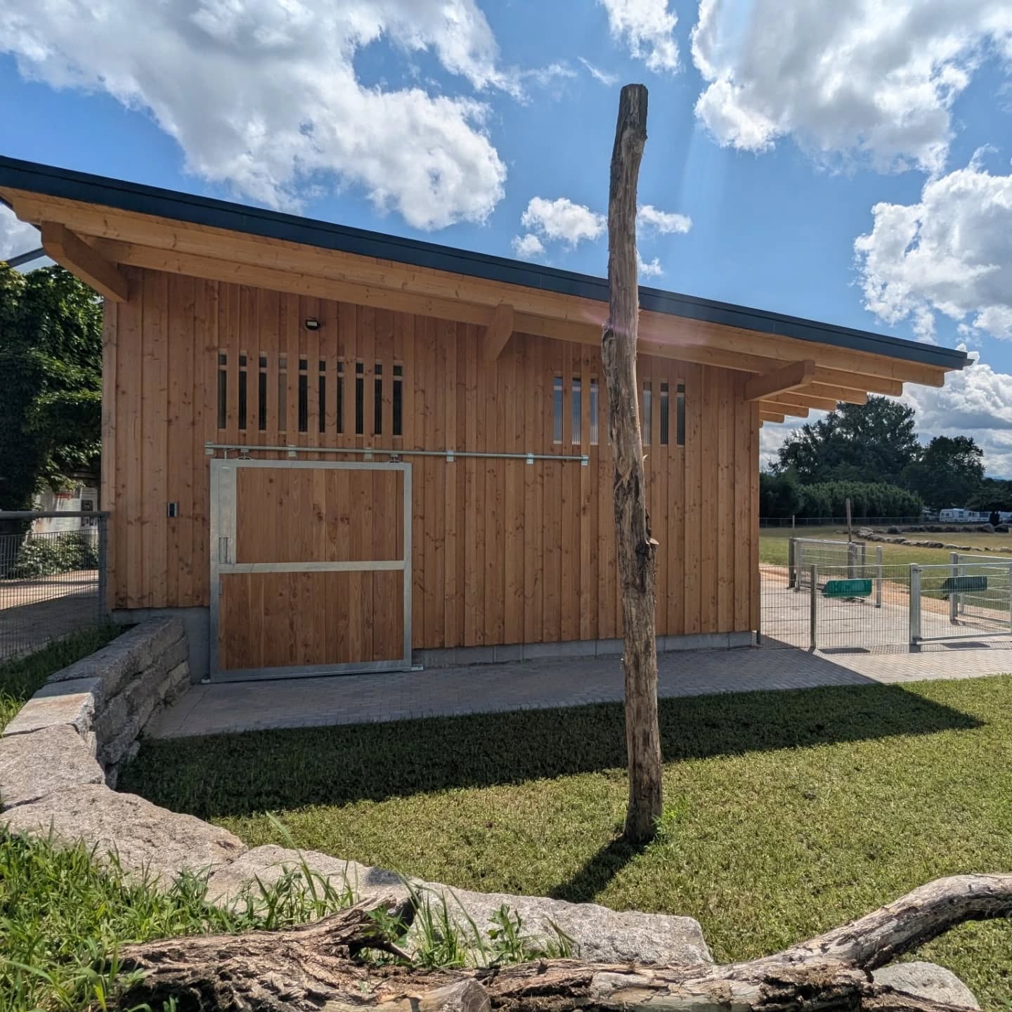 Das Foto zeigt ein Holzgebäude mit einem grünen Rasen im Vordergrund unter einem blauen Himmel mit Wolken.