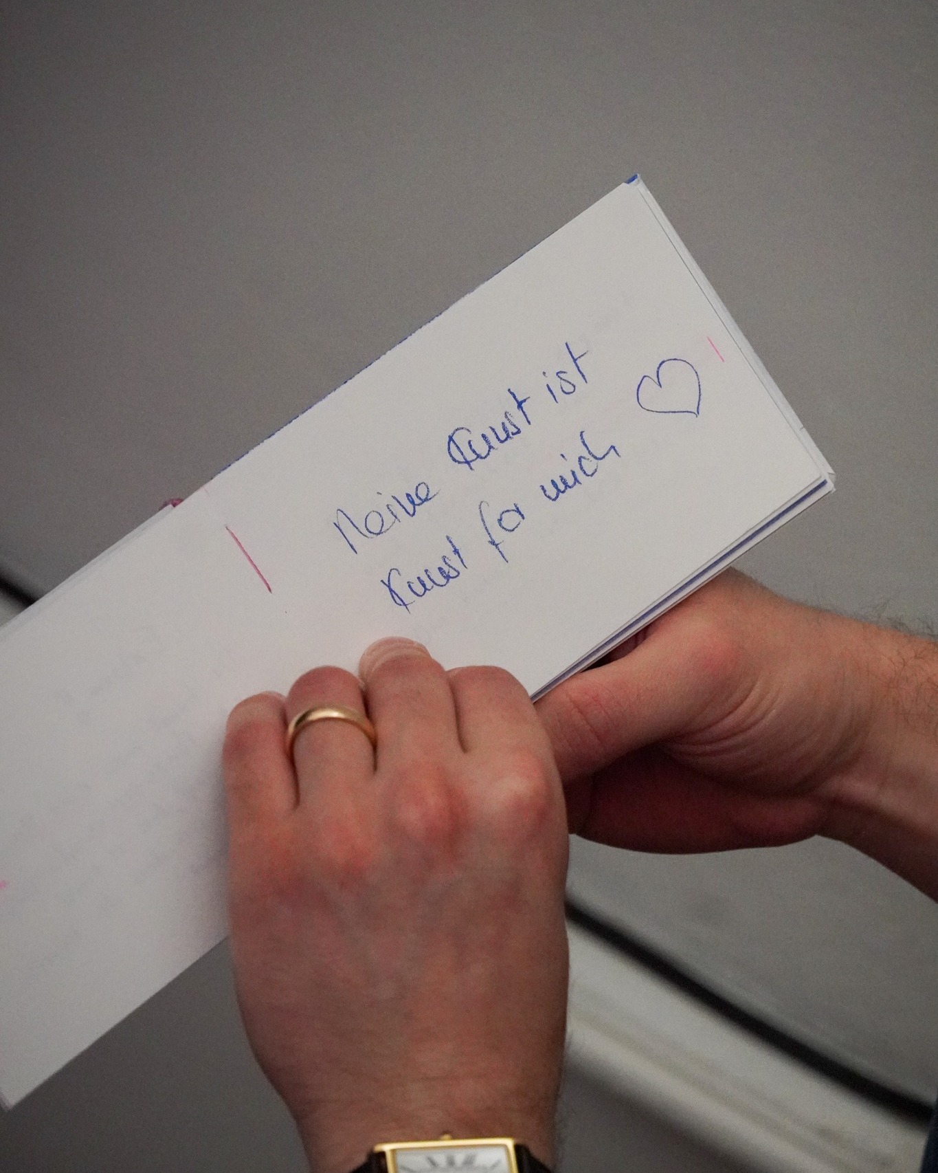 Dieser Alternativtext wurde KI-generiert: Eine Hand hält einen gefalteten Zettel mit handgeschriebener Schrift und einer Zeichnung. Ein goldener Ring ist an einem Finger zu sehen.