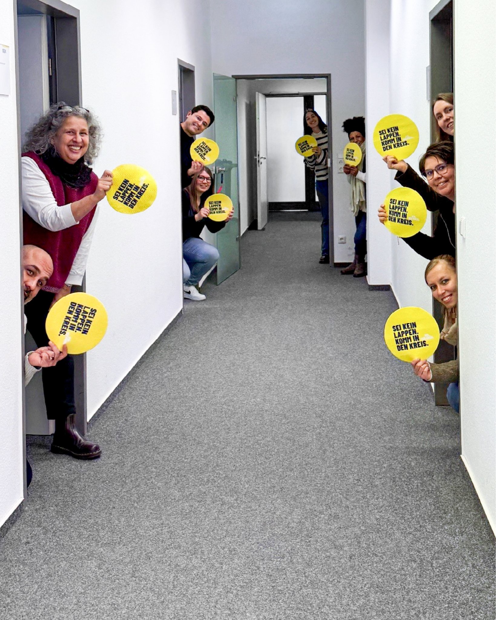 Dieser Alternativtext wurde KI-generiert: Eine Gruppe von Personen hält Schilder mit Aufschriften hoch. Das farbige Bild zeigt sie in einem Flur stehend.