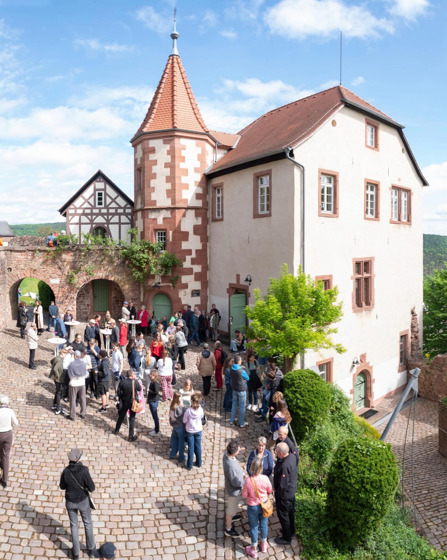 Dieser Alternativtext wurde KI-generiert: Ein historisches Gebäude mit einem roten Dach und einem markanten Turm ist im Bild zu sehen. Vor dem Gebäude befindet sich eine Gruppe von Menschen auf einem gepflasterten Platz.