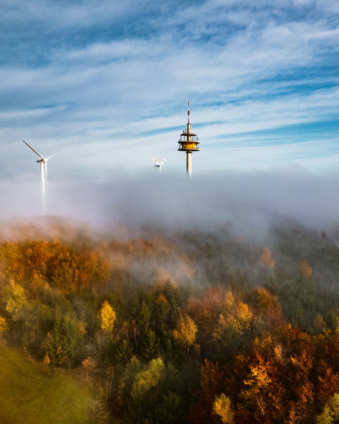 Dieser Alternativtext wurde KI-generiert: Eine Aufnahme zeigt einen Turm und Windräder über einer Nebeldecke. Im Vordergrund sind Bäume mit Herbstlaub zu sehen.