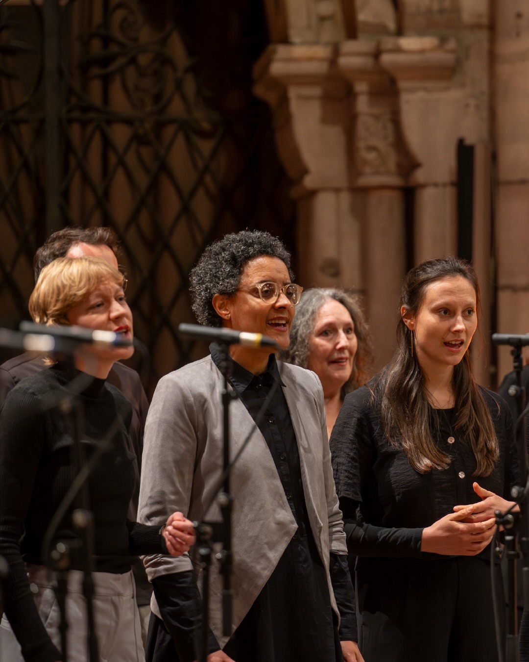 Dieser Alternativtext wurde KI-generiert: Eine Gruppe von Menschen steht vor einer verzierten Wand und singt oder spricht in Mikrofone. Das Bild ist farbig und zeigt eine Innenraumansicht.