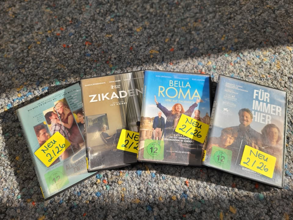 Dieser Alternativtext wurde KI-generiert: Mehrere DVD-Hüllen liegen nebeneinander auf einem bunten Teppich. Einige Hüllen tragen gelbe Aufkleber mit Zahlen.